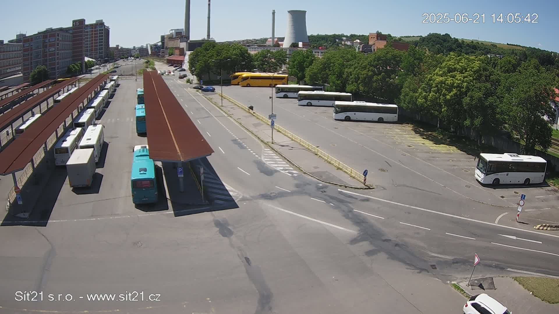A bus station with numerous buses parked under covered areas and in open lots on a sunny day, next to industrial buildings and green trees.