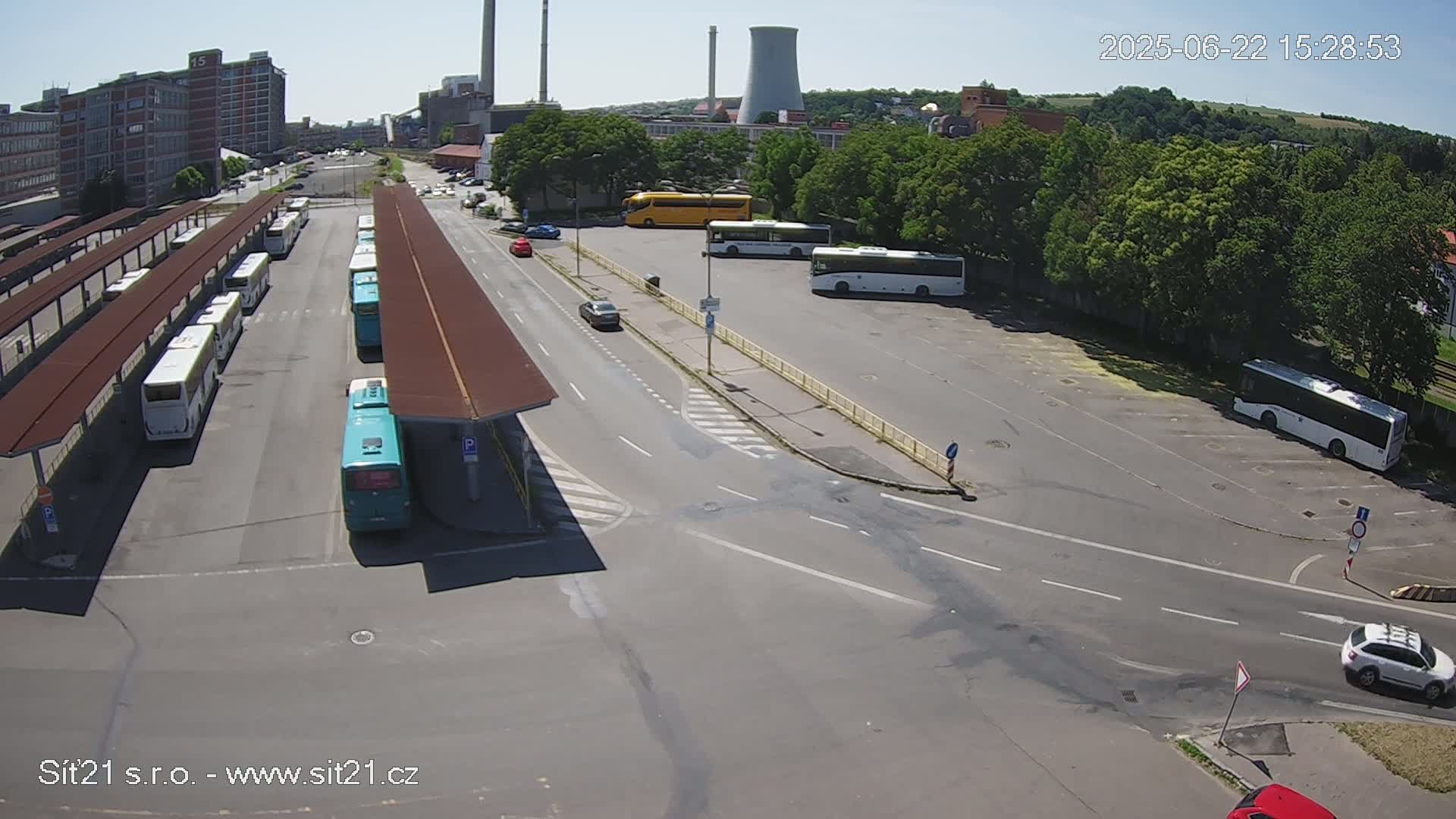 A bus station with numerous buses parked under shelters, cars driving on the roads surrounding it, and industrial buildings and trees visible in the background on a sunny day.