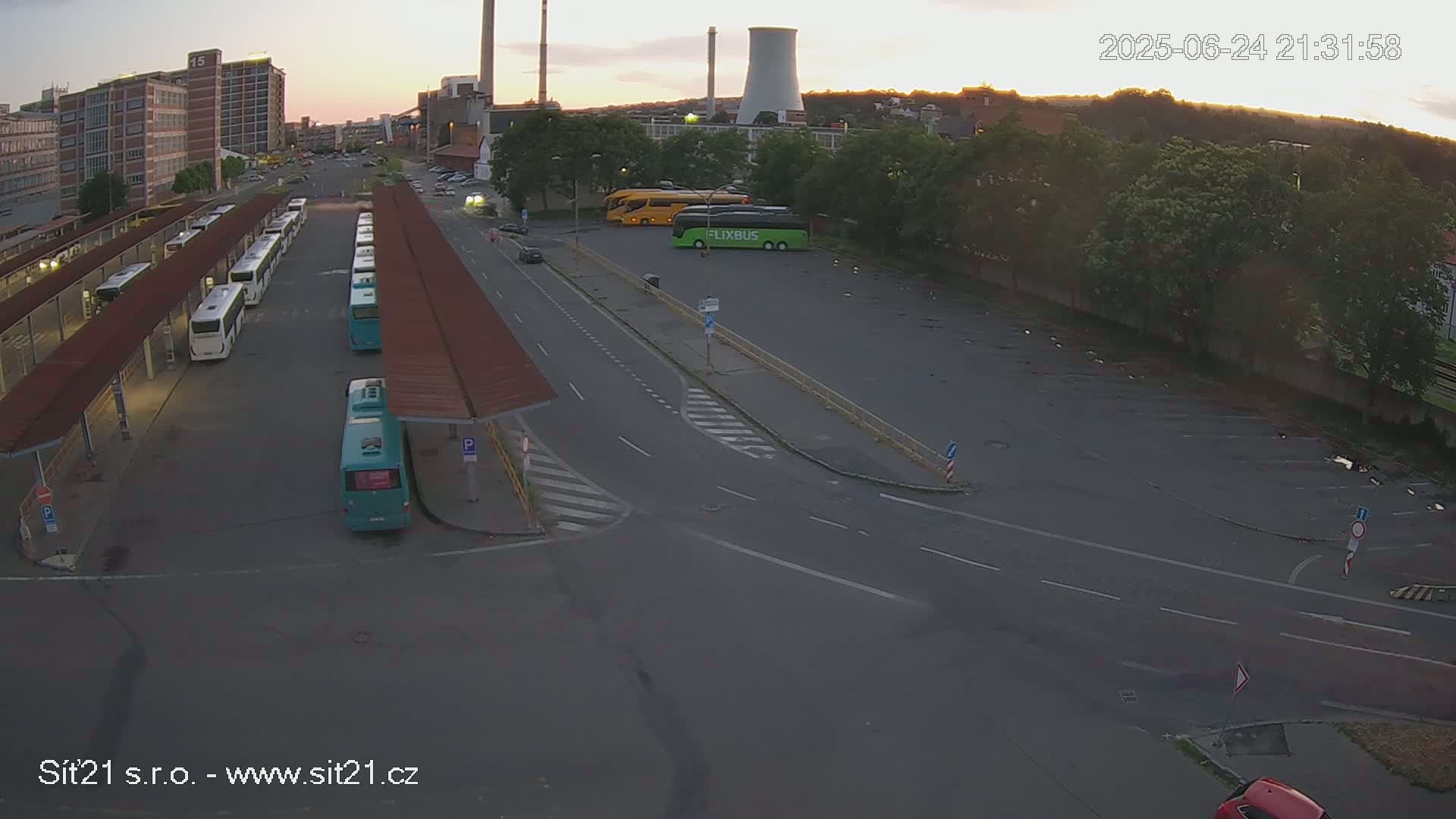 A high-angle, twilight view shows a bus station with numerous buses parked under covered areas, along with additional buses and cars in a nearby parking lot, adjacent to an industrial area.