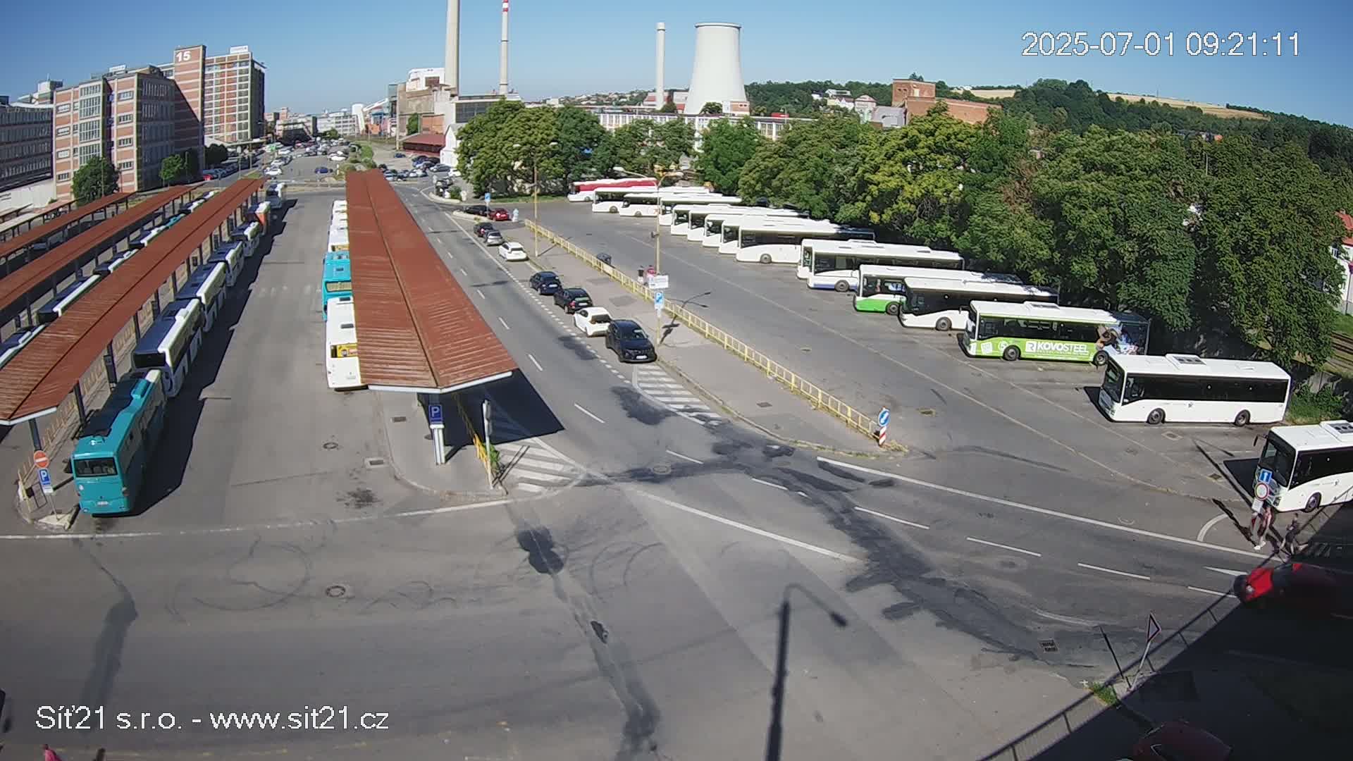 A bus station with numerous buses parked under covered shelters and in an adjacent open lot on a sunny day.