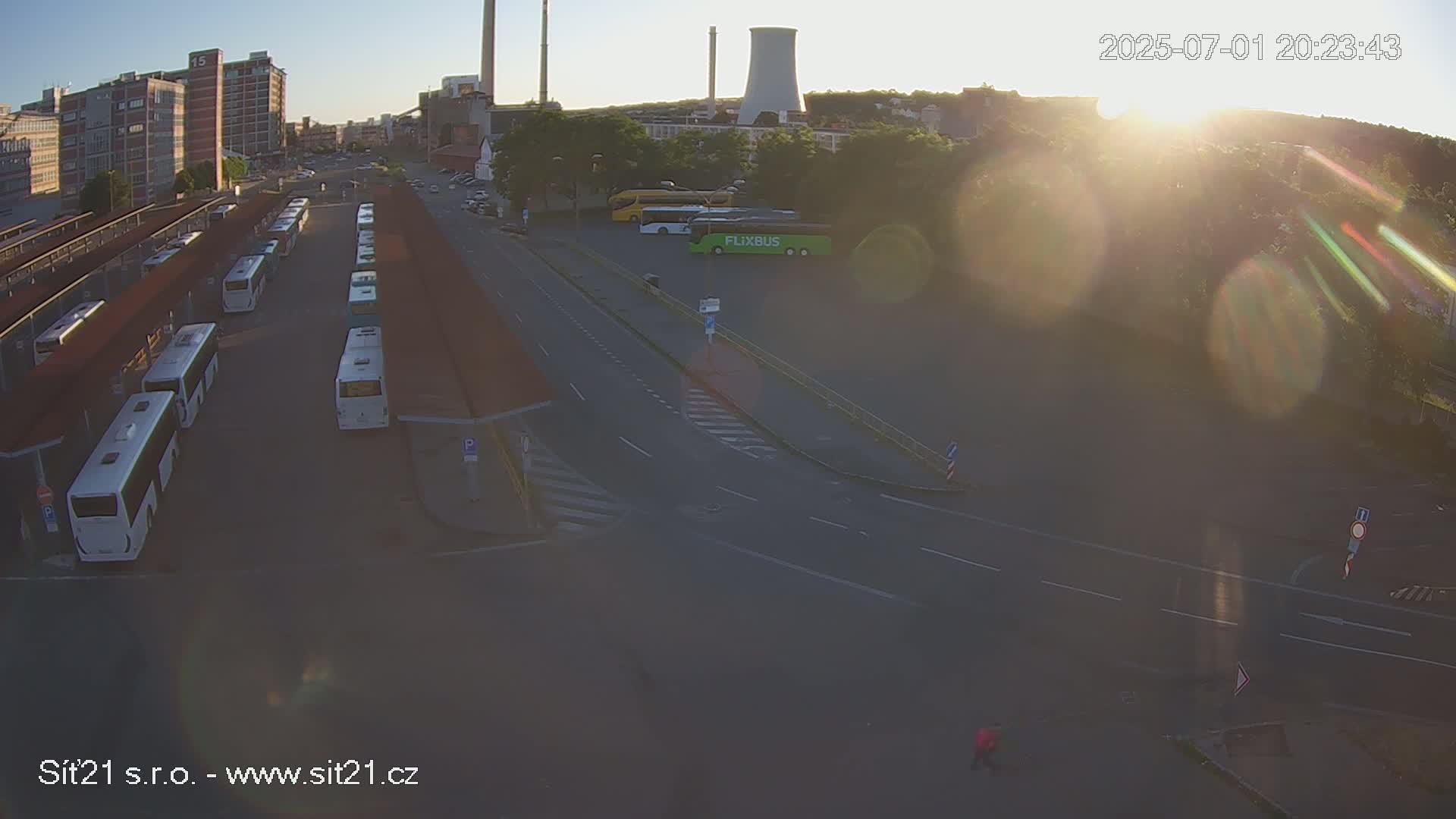 A high-angle, long shot shows a bus station with numerous buses parked under covered shelters, next to a road intersection under a hazy, bright sunrise.