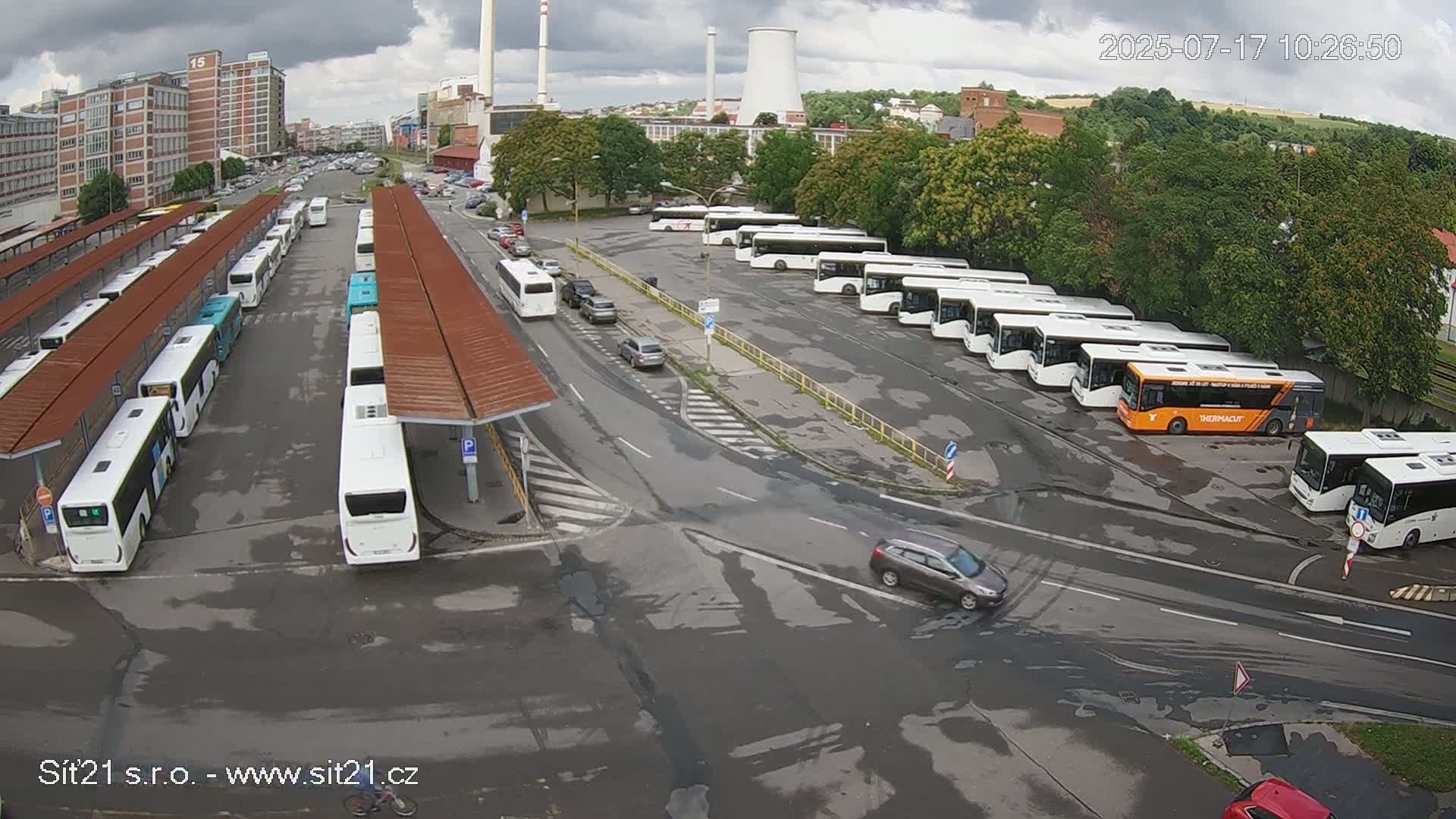 Many buses are parked at a bus station under an overcast sky, with a few cars driving on nearby roads.