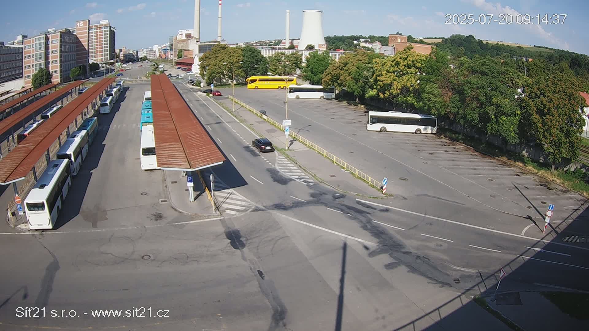 A sunny day at a bus station with several buses parked under shelters, a few cars in a parking lot, and industrial buildings in the background.