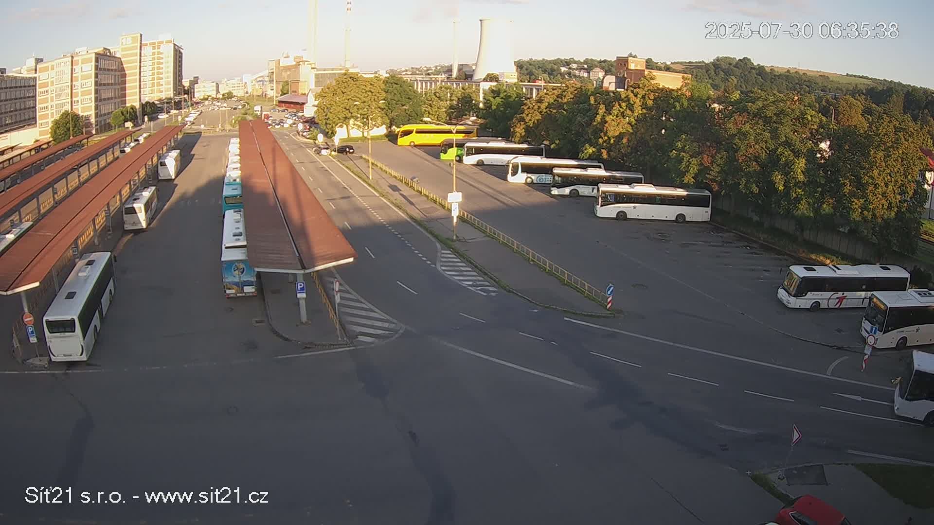 A sunny day at a bus station with several buses parked under shelters, a few cars in a parking lot, and industrial buildings in the background.