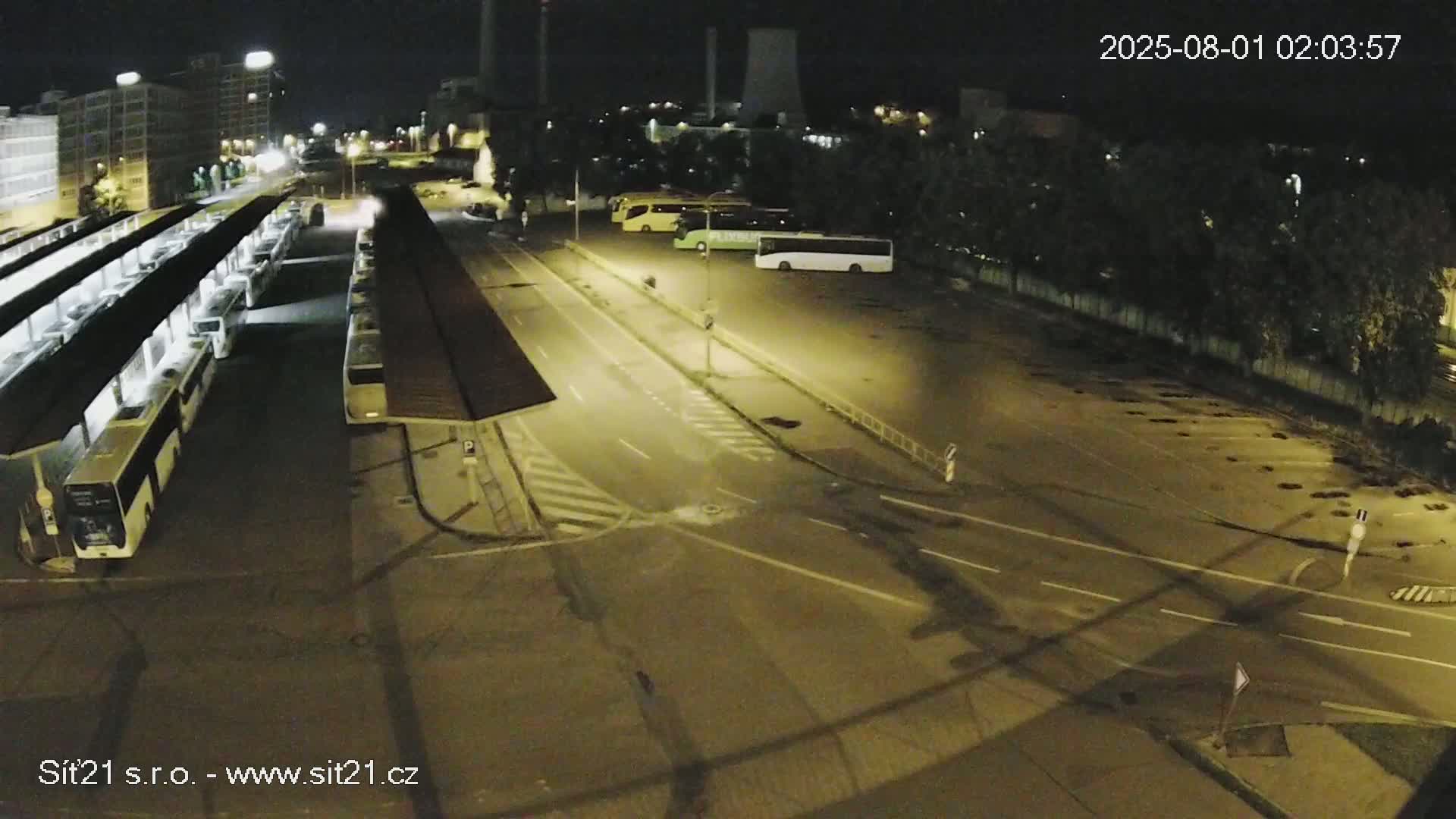 At night, a bus station with numerous buses parked under shelters and a few in a nearby lot is illuminated.