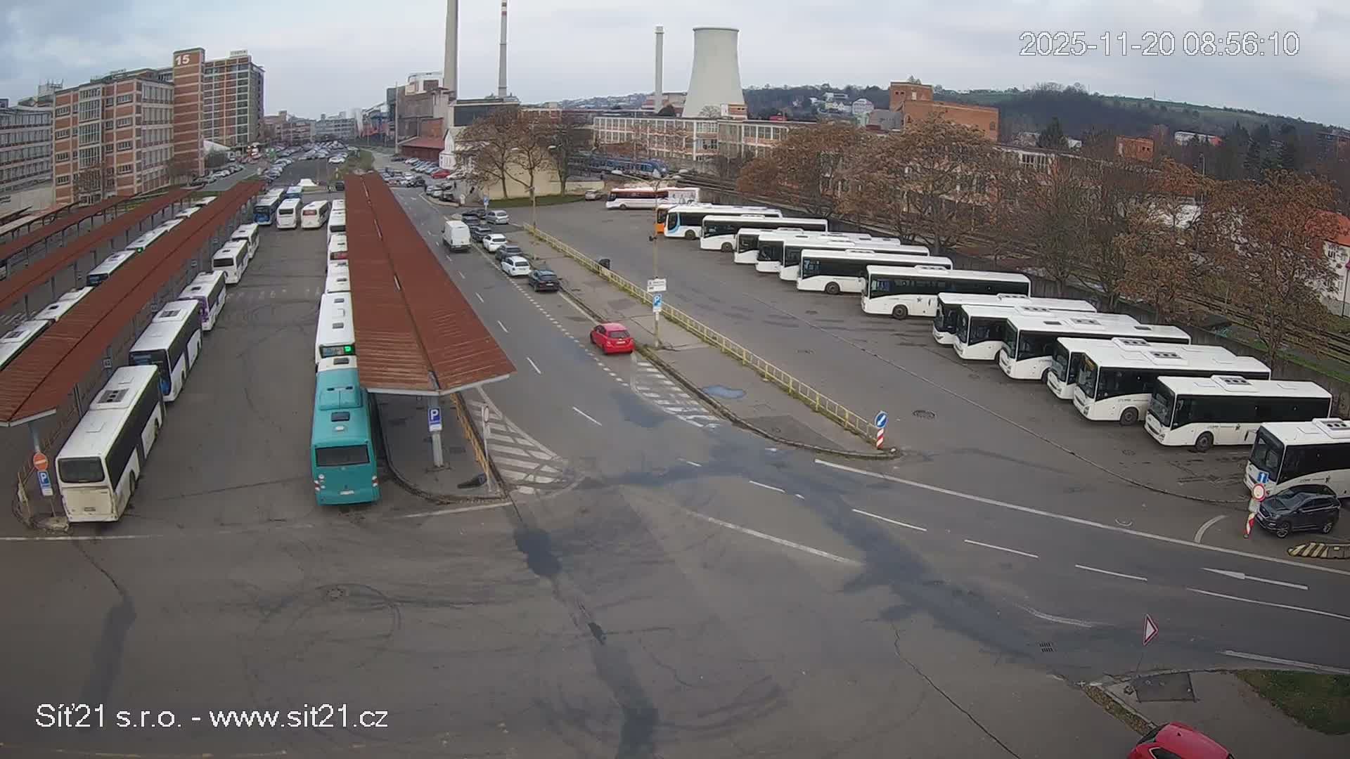 An elevated view shows an extensive outdoor bus terminal filled with many parked white and one turquoise bus, alongside roads with various vehicles and a distant backdrop of industrial structures, urban buildings, and autumn trees, all beneath a cloudy, grey sky.