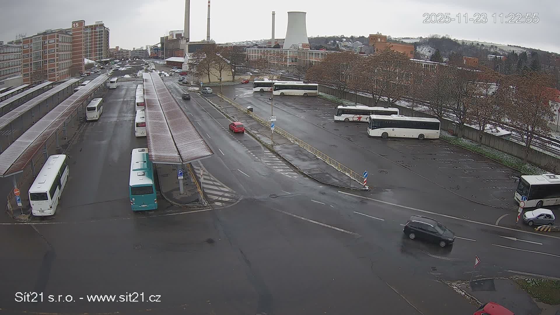 An overcast sky hangs over a city scene featuring a large bus depot with numerous buses, wet roads, and industrial structures, with distant hills showing patches of snow.