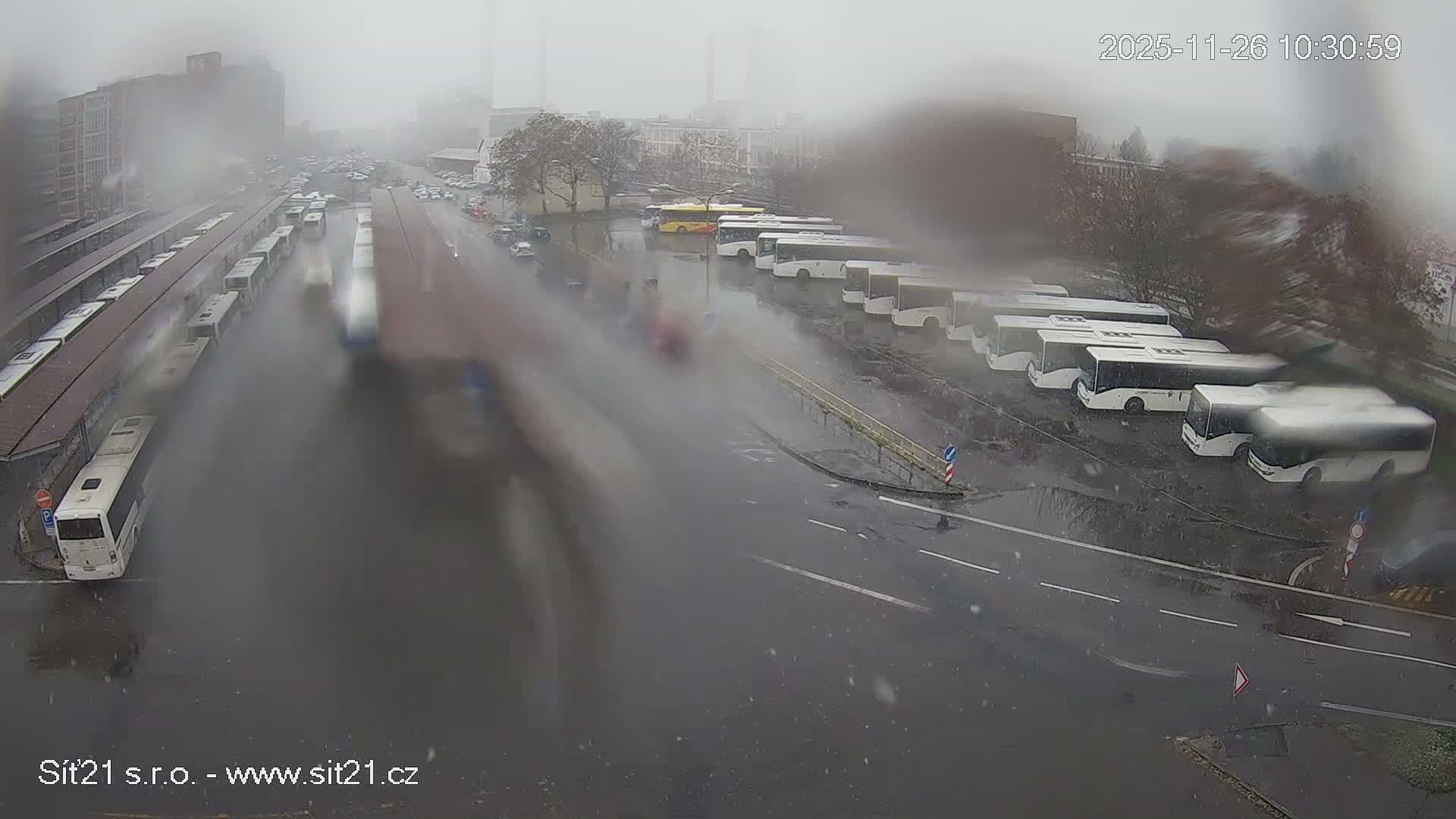 A blurry, high-angle view captures a busy outdoor bus terminal and adjacent streets filled with parked and moving buses and cars on a wet and heavily precipitating day, suggesting rain or snow.