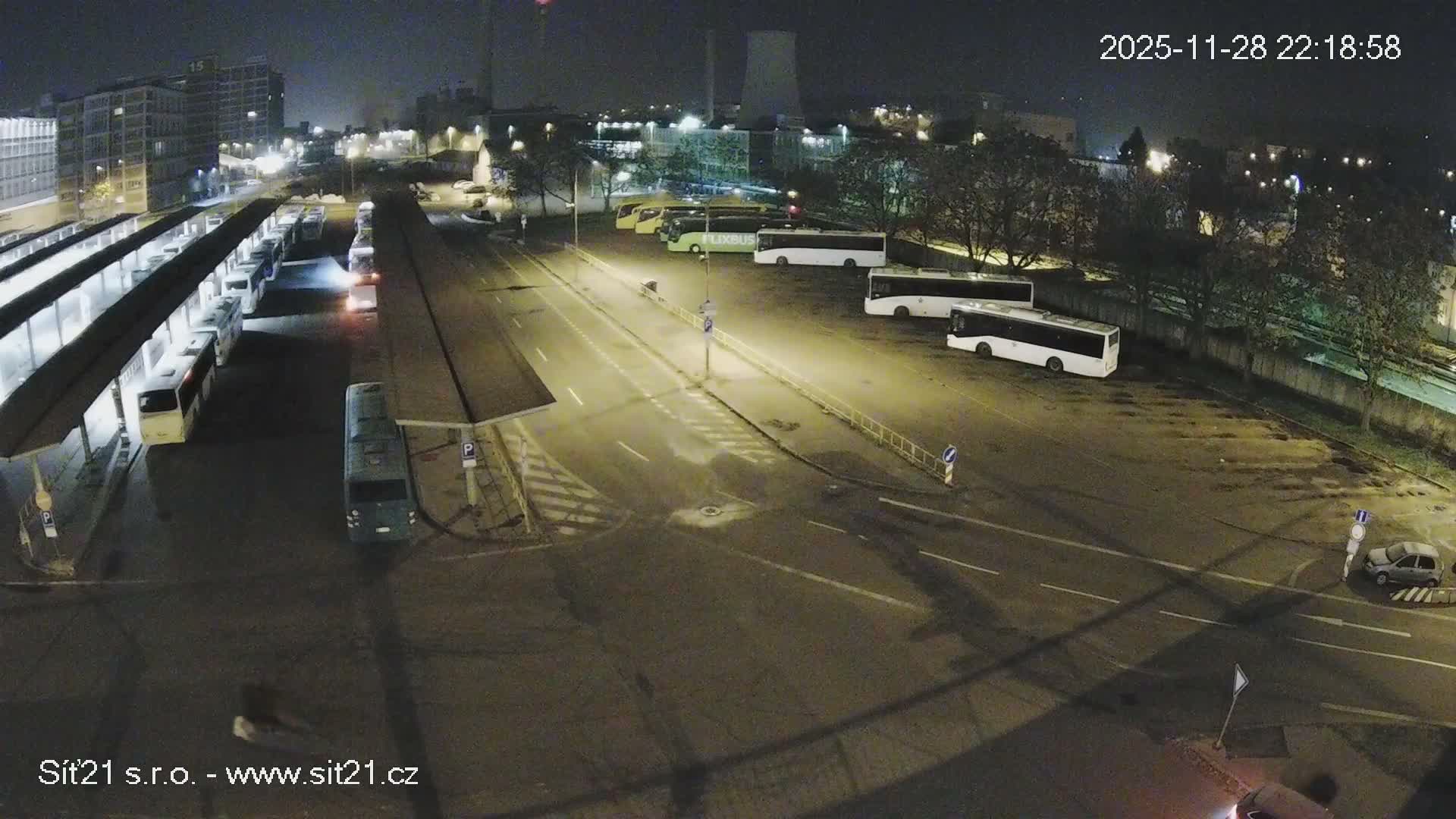 The image captures an expansive, brightly lit bus terminal and adjacent street at night under clear skies, with numerous buses parked at platforms and in a lot, and buildings visible in the distance.