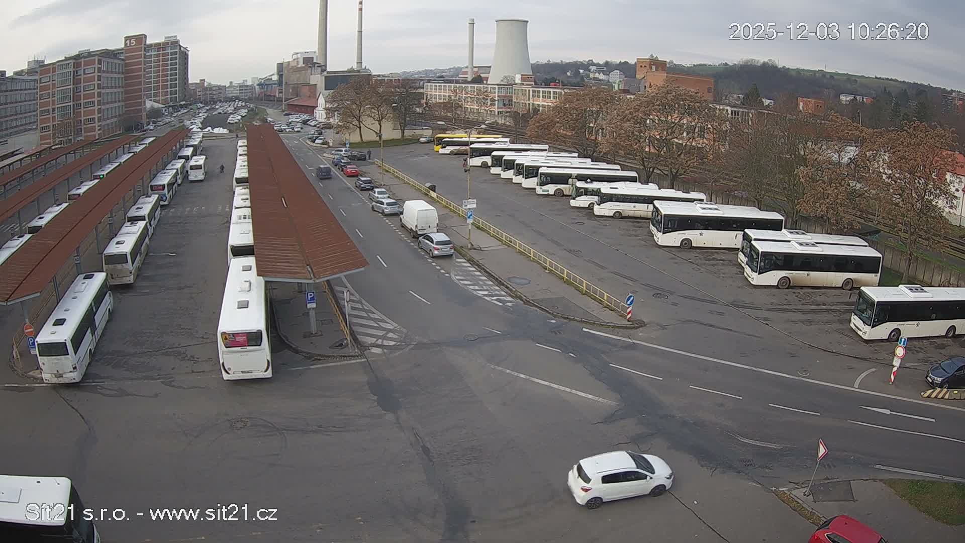 Numerous white buses are parked at an outdoor station with shelters, alongside a road with cars, set against an urban backdrop featuring industrial buildings and a cooling tower under an overcast sky.