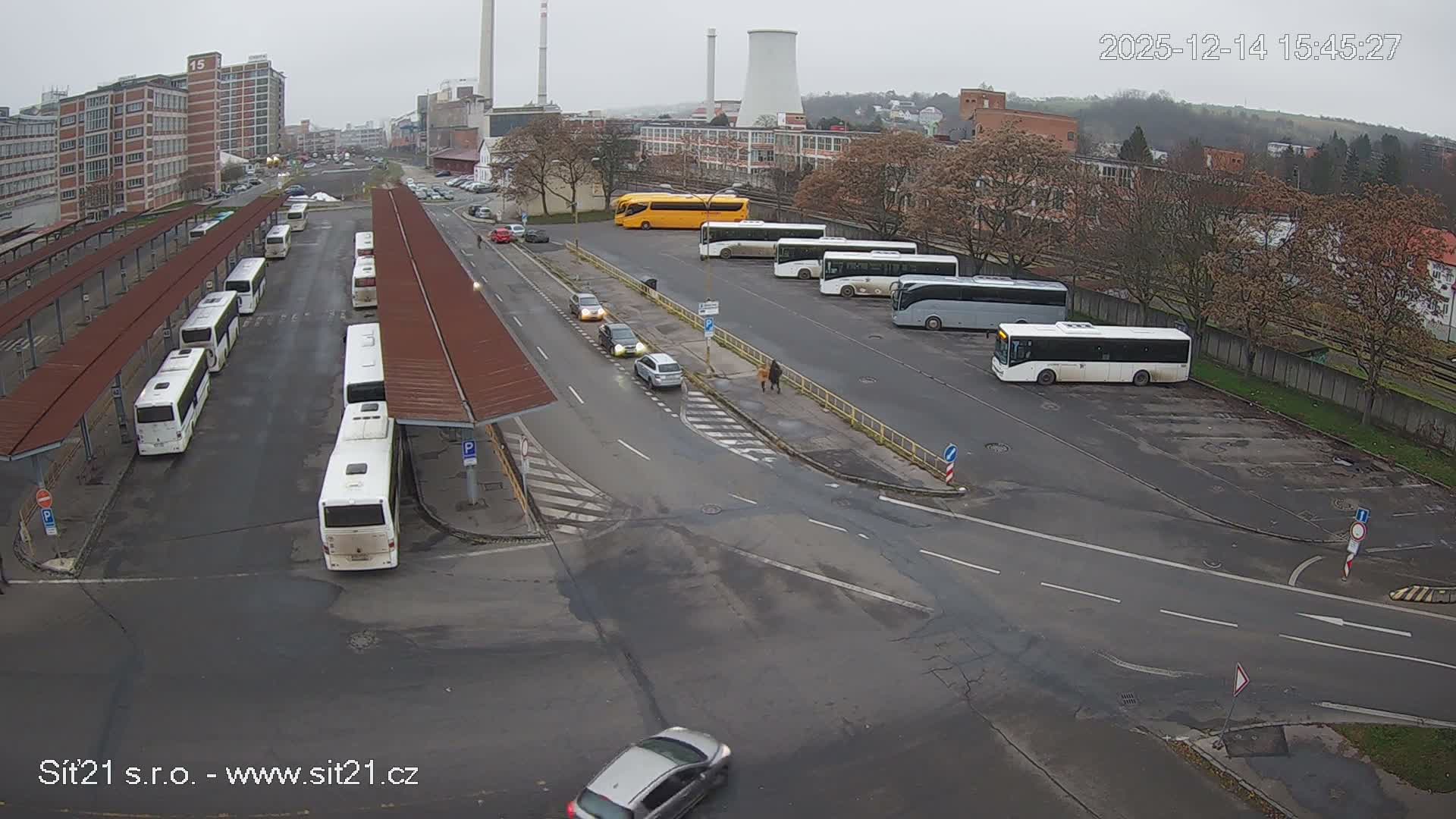 Numerous white buses are parked at an outdoor station with shelters, alongside a road with cars, set against an urban backdrop featuring industrial buildings and a cooling tower under an overcast sky.