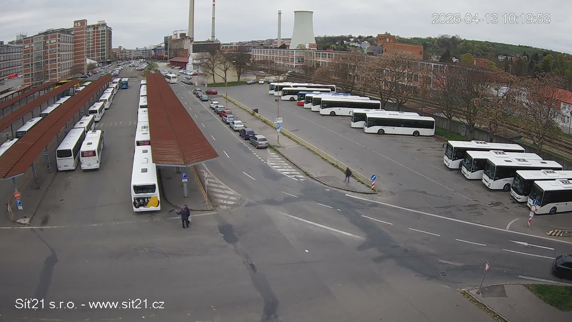 Numerous white buses are parked at an outdoor station with shelters, alongside a road with cars, set against an urban backdrop featuring industrial buildings and a cooling tower under an overcast sky.