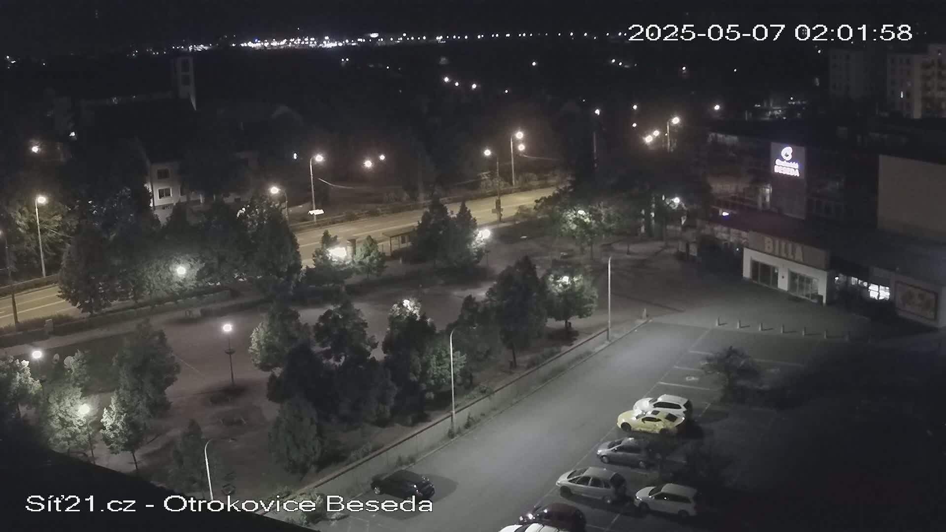 A nighttime, overhead view of a parking lot with several cars, surrounded by trees and streetlights, next to a roadway.