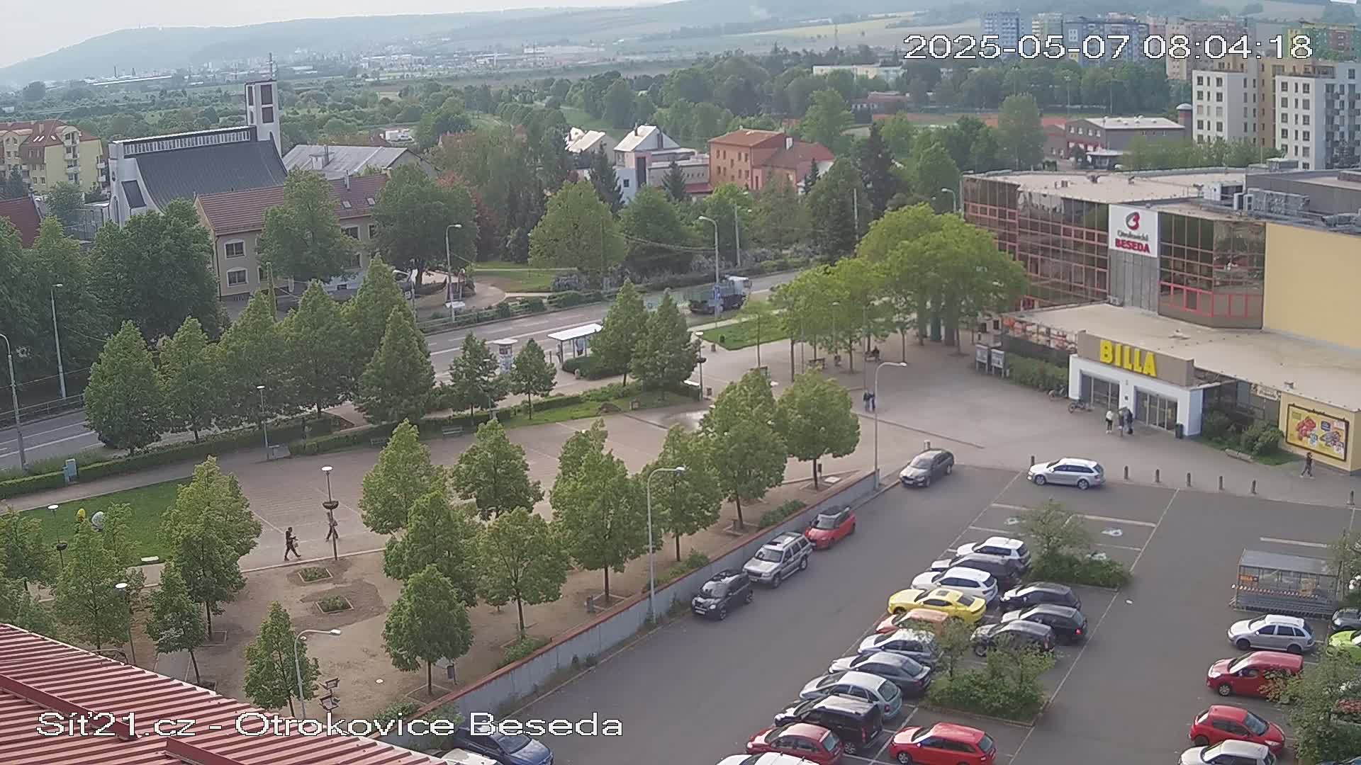 An aerial view shows a town square with a church, a supermarket, and a parking lot filled with cars under an overcast sky.