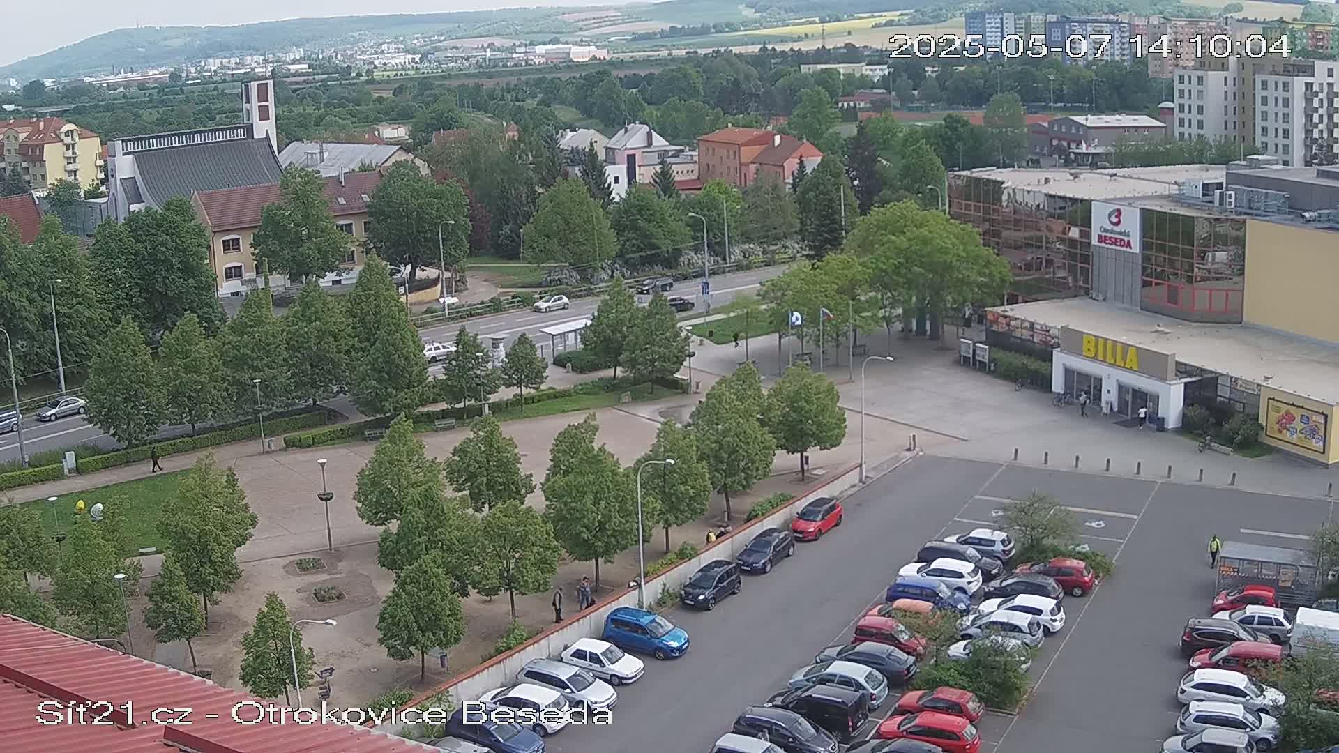 A high-angle view shows a town square with a church, a supermarket, and a parking lot filled with cars under a mostly cloudy sky.