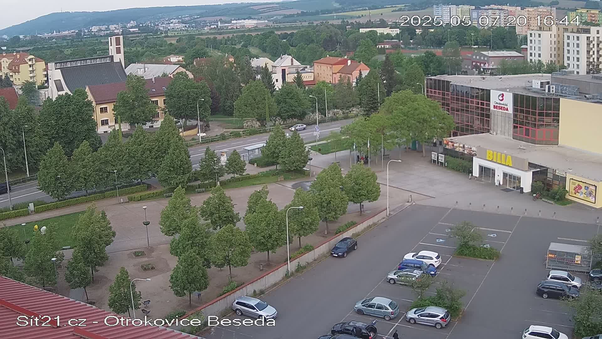 An aerial view of a town square with a church, a large building, and a parking lot, under a mostly clear sky.