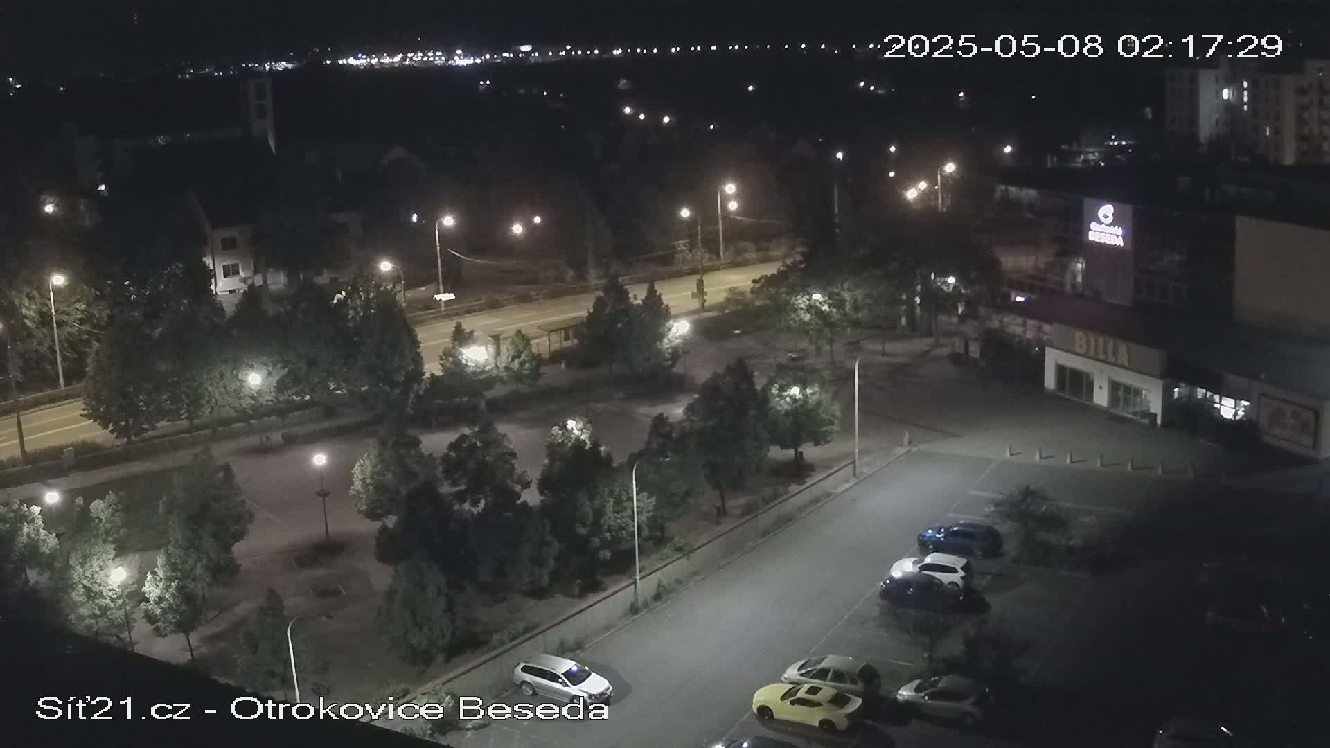A nighttime, overhead view of a parking lot with several cars, surrounded by trees and streetlights, next to a road and buildings.