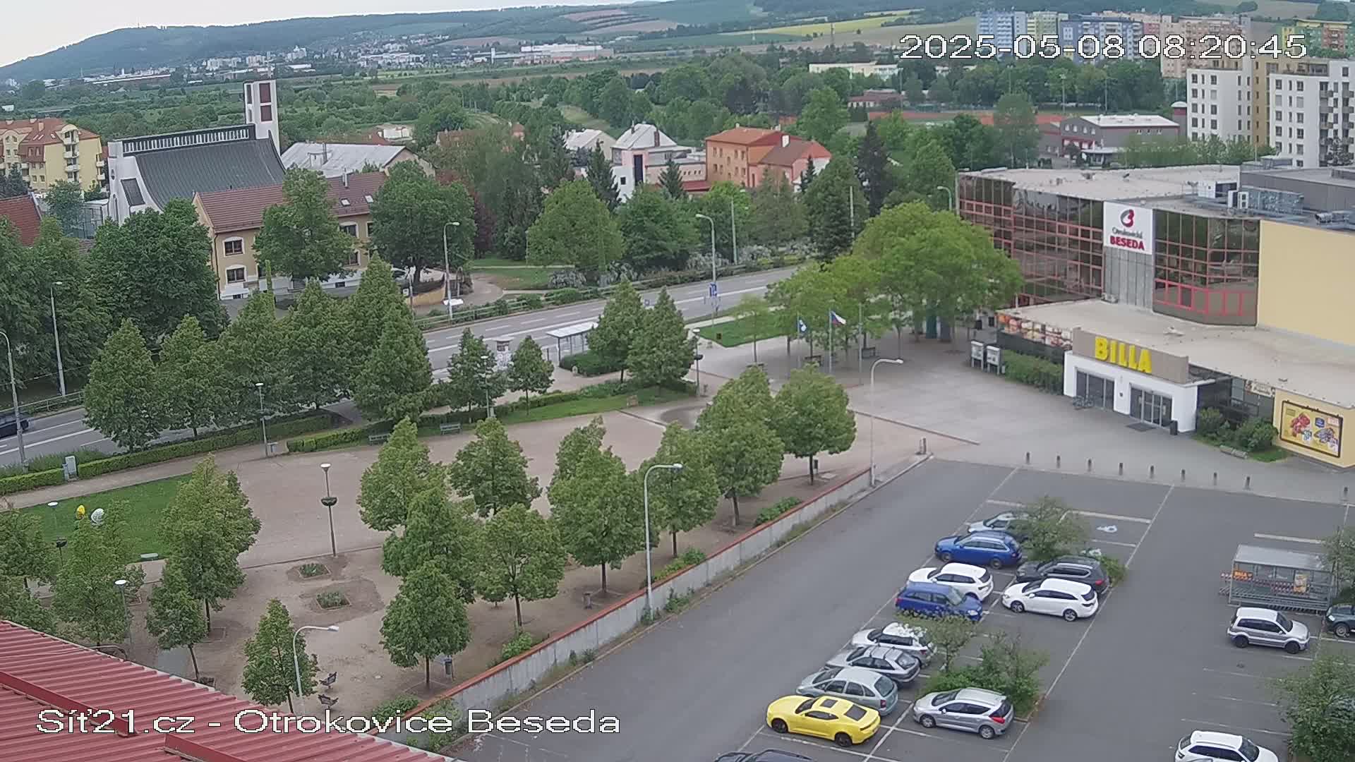 An aerial view shows a town square with green trees, a church, a large building, and a parking lot filled with cars on an overcast day.