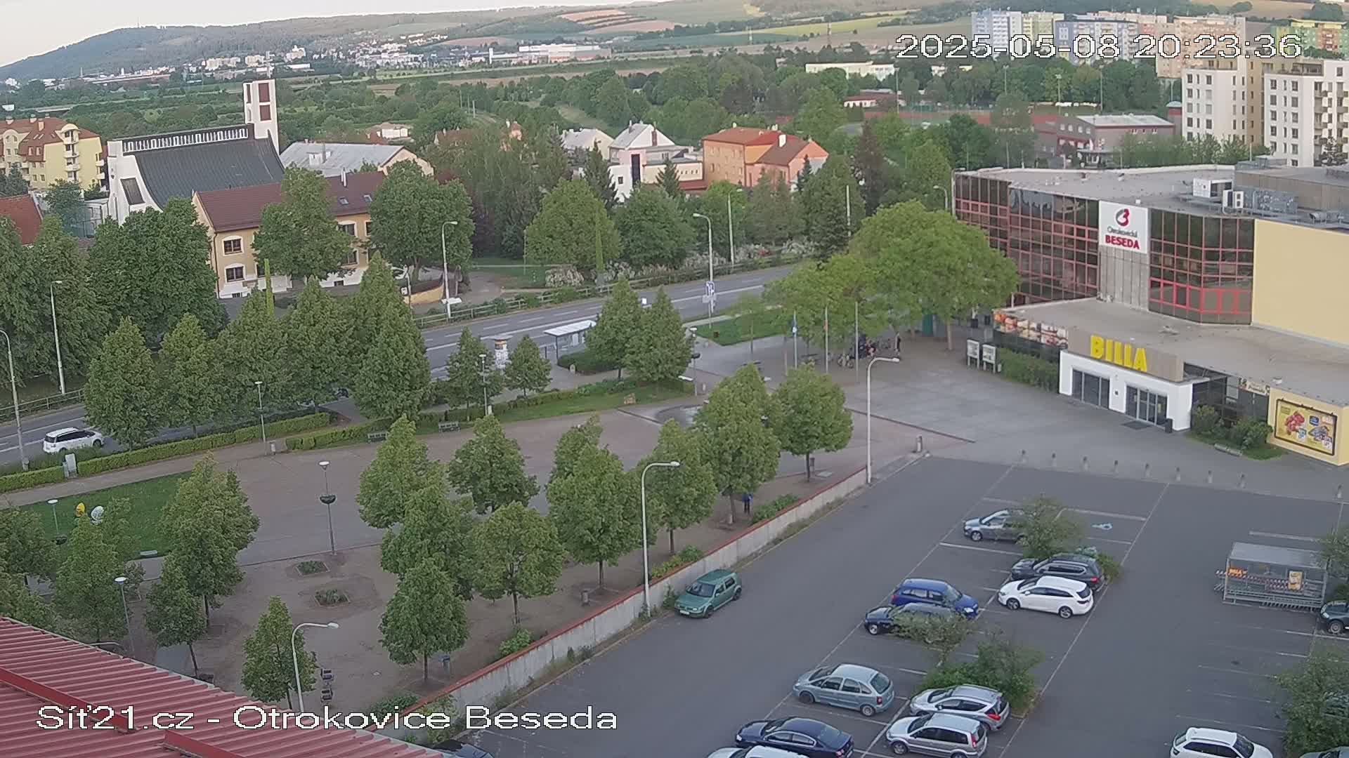 An elevated view of a town square with a church, buildings, a parking lot, and green trees under a partly cloudy sky.