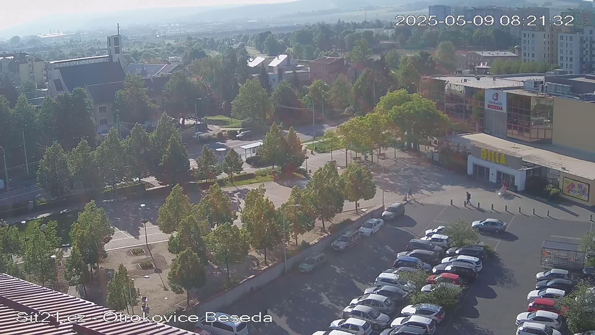 An aerial view of a town square with a church, shops, a parking lot filled with cars, and many trees under a hazy, sunny sky.