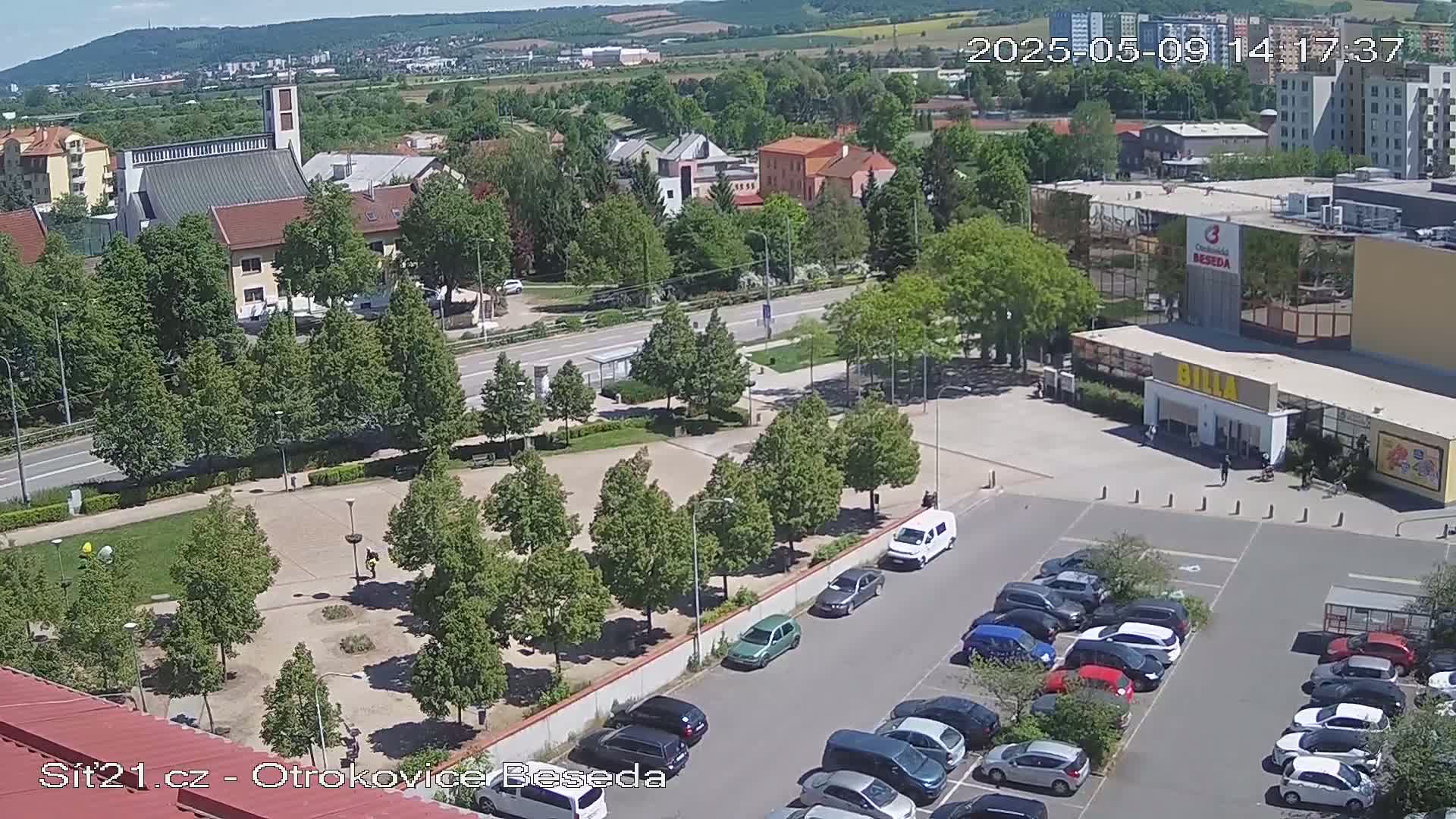 An elevated view shows a sunny day overlooking a town square with a church, buildings, a parking lot full of cars, and green trees.
