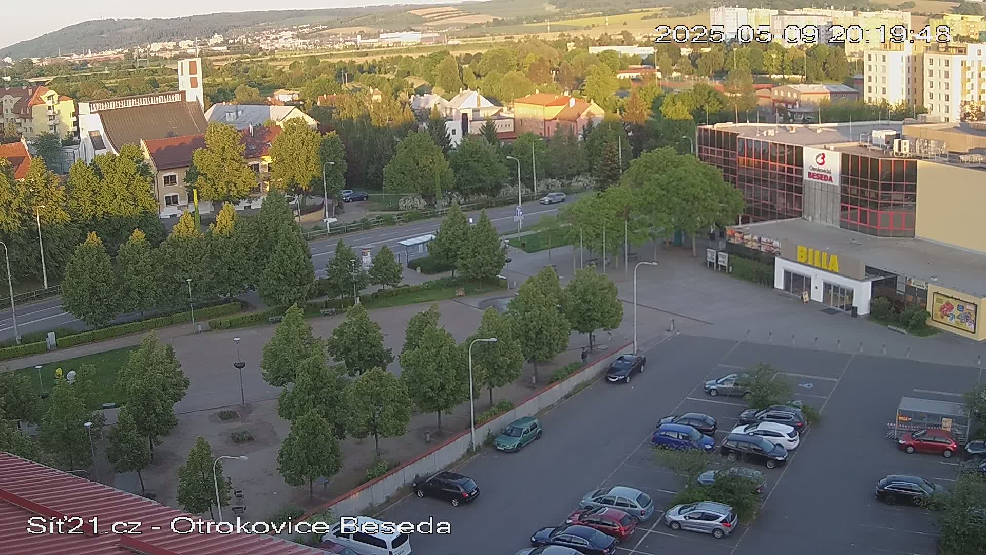 An elevated view of a town square at sunset, showing a church, buildings, a parking lot with cars, and numerous green trees lining the streets and plaza.