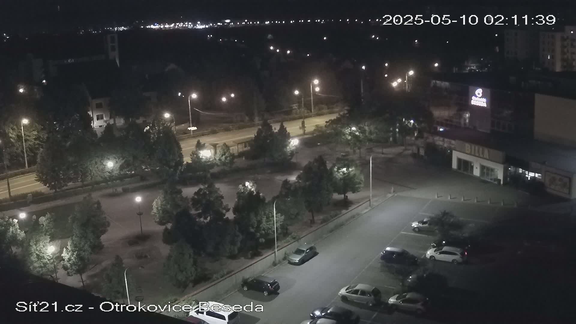 A nighttime aerial view of a parking lot with several cars, surrounded by trees and streetlights, next to a road and buildings.