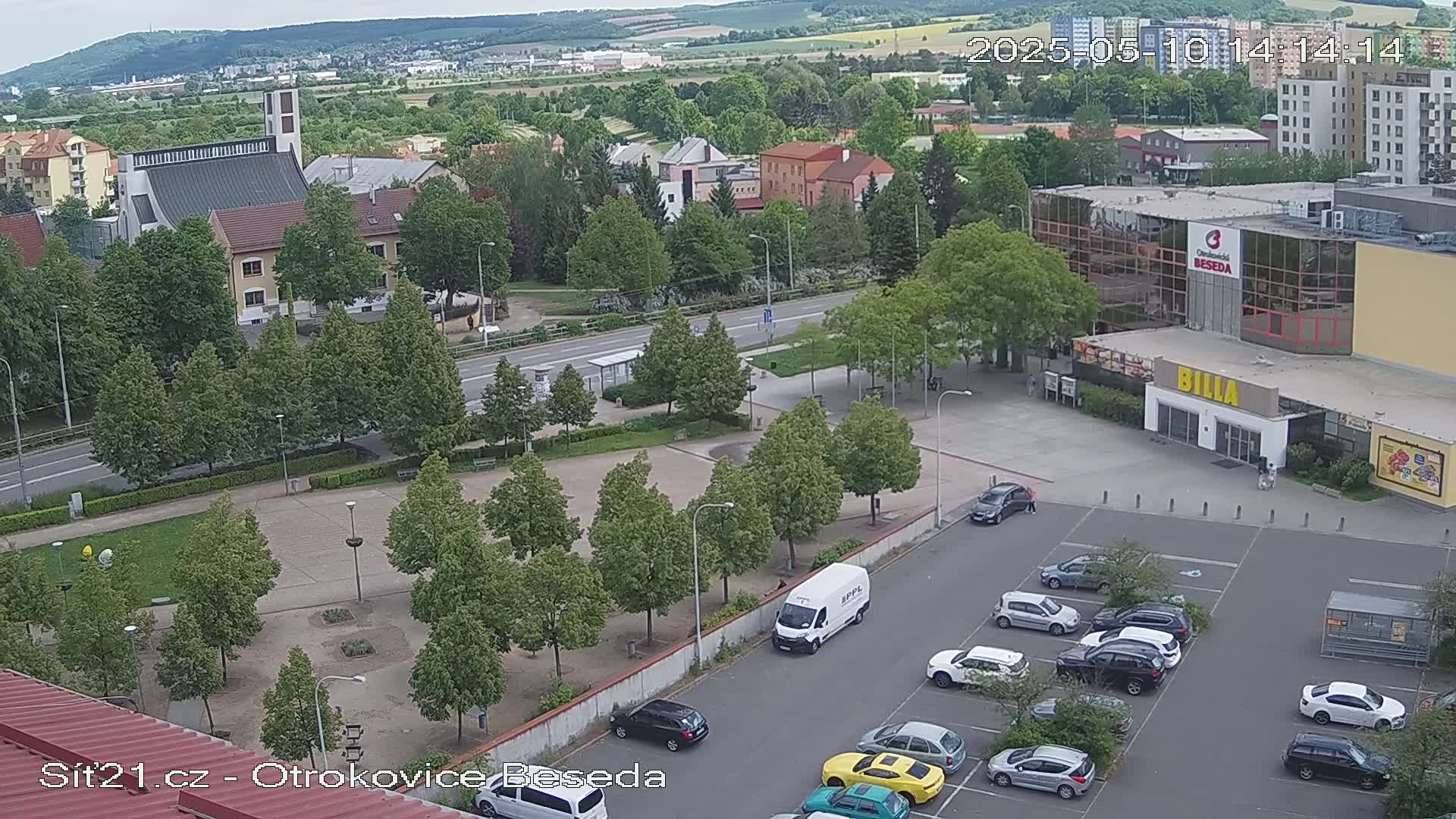 An elevated view of a town square with a church, a large building, a parking lot full of cars, and many green trees on a partly cloudy day.