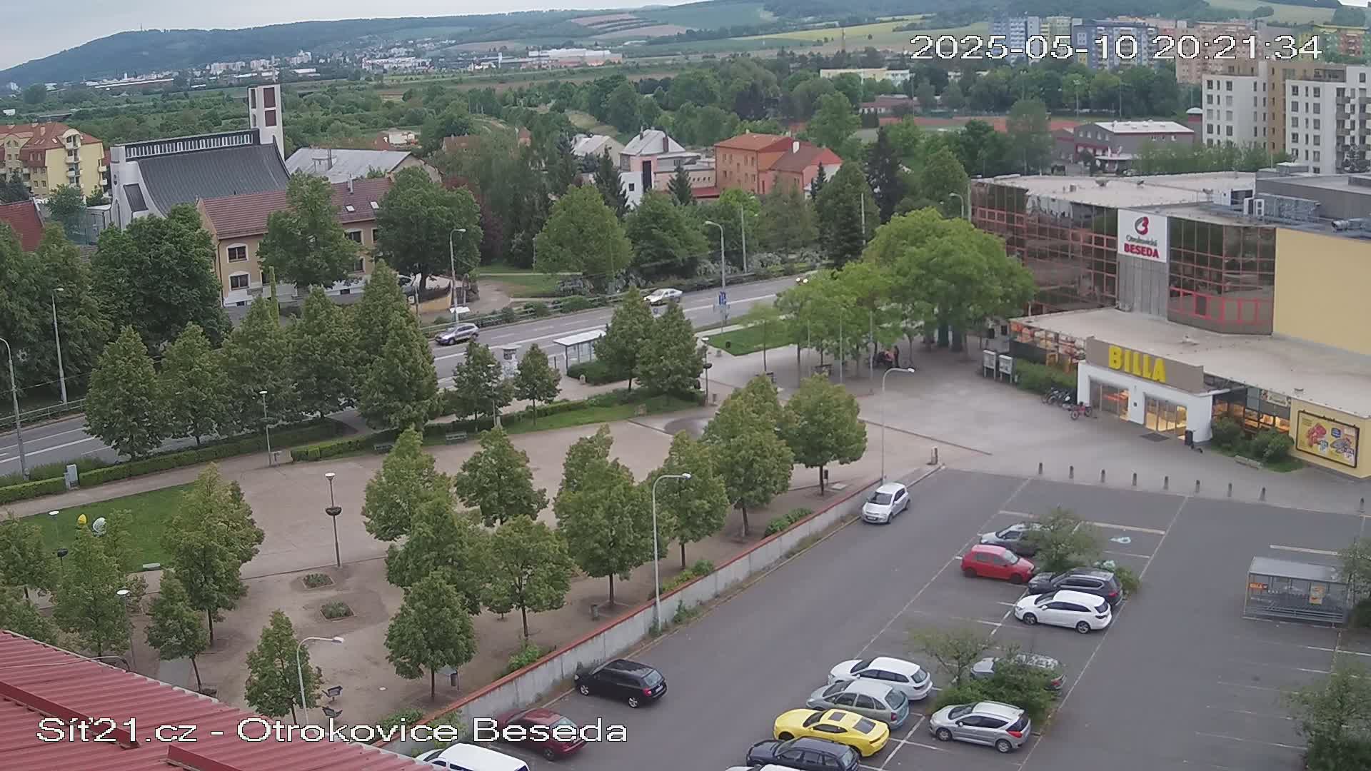 An elevated view of a town square with a church, buildings, a supermarket, and a parking lot under an overcast sky.