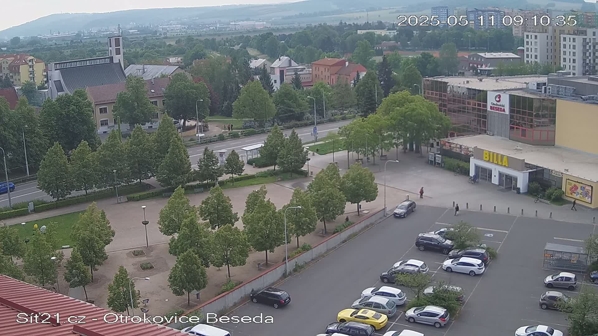 An elevated view shows a town square with a church, buildings, a supermarket, and a parking lot on an overcast day.