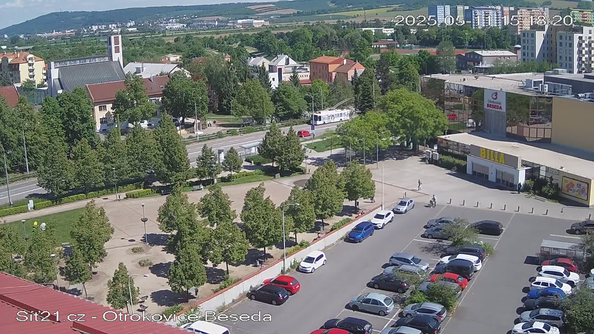 An aerial view shows a town square with a church, a supermarket, and a parking lot full of cars on a sunny day.