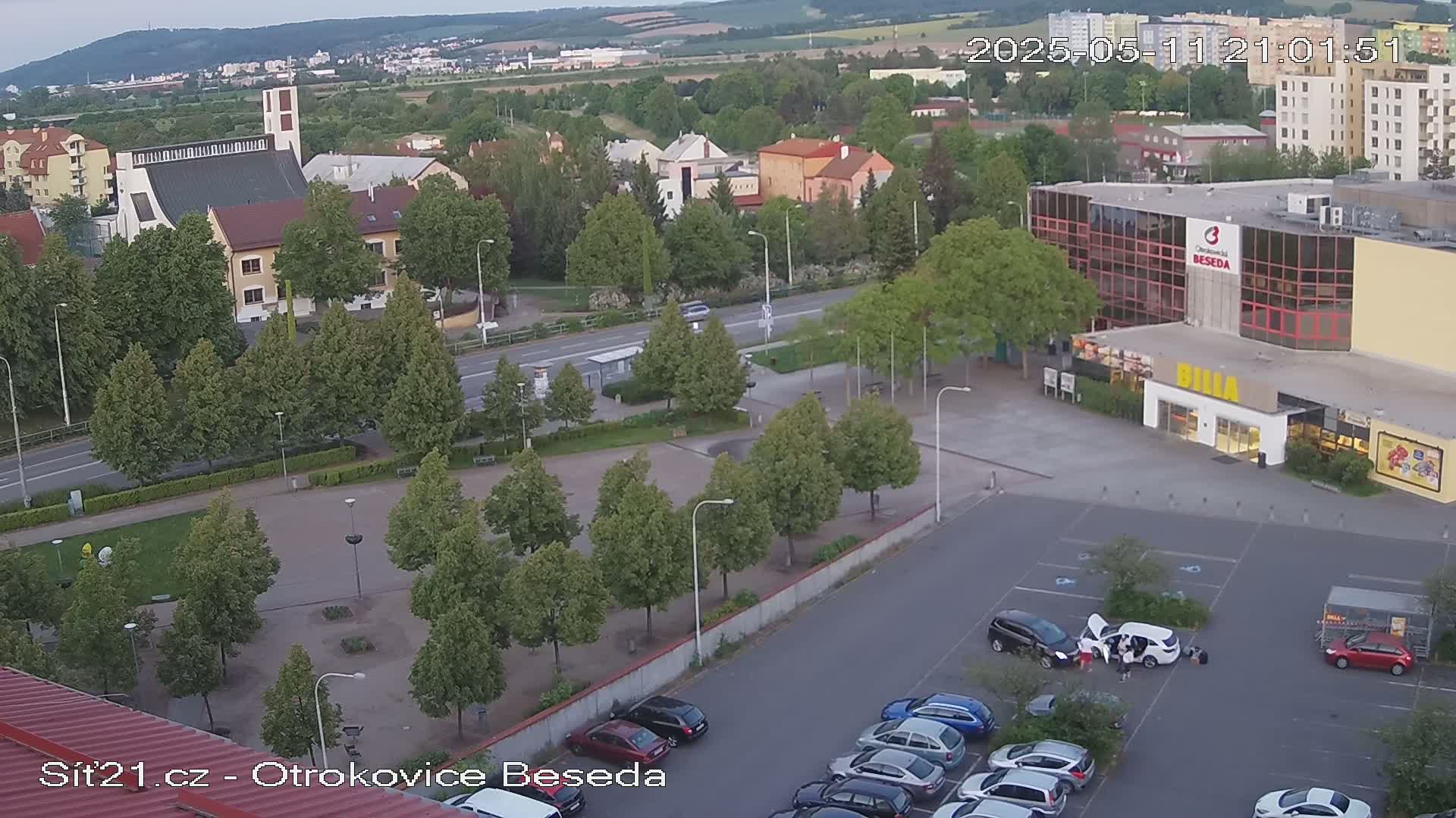 An aerial view shows a town square with a church, several buildings, a parking lot with cars, and green trees under a partly cloudy sky.