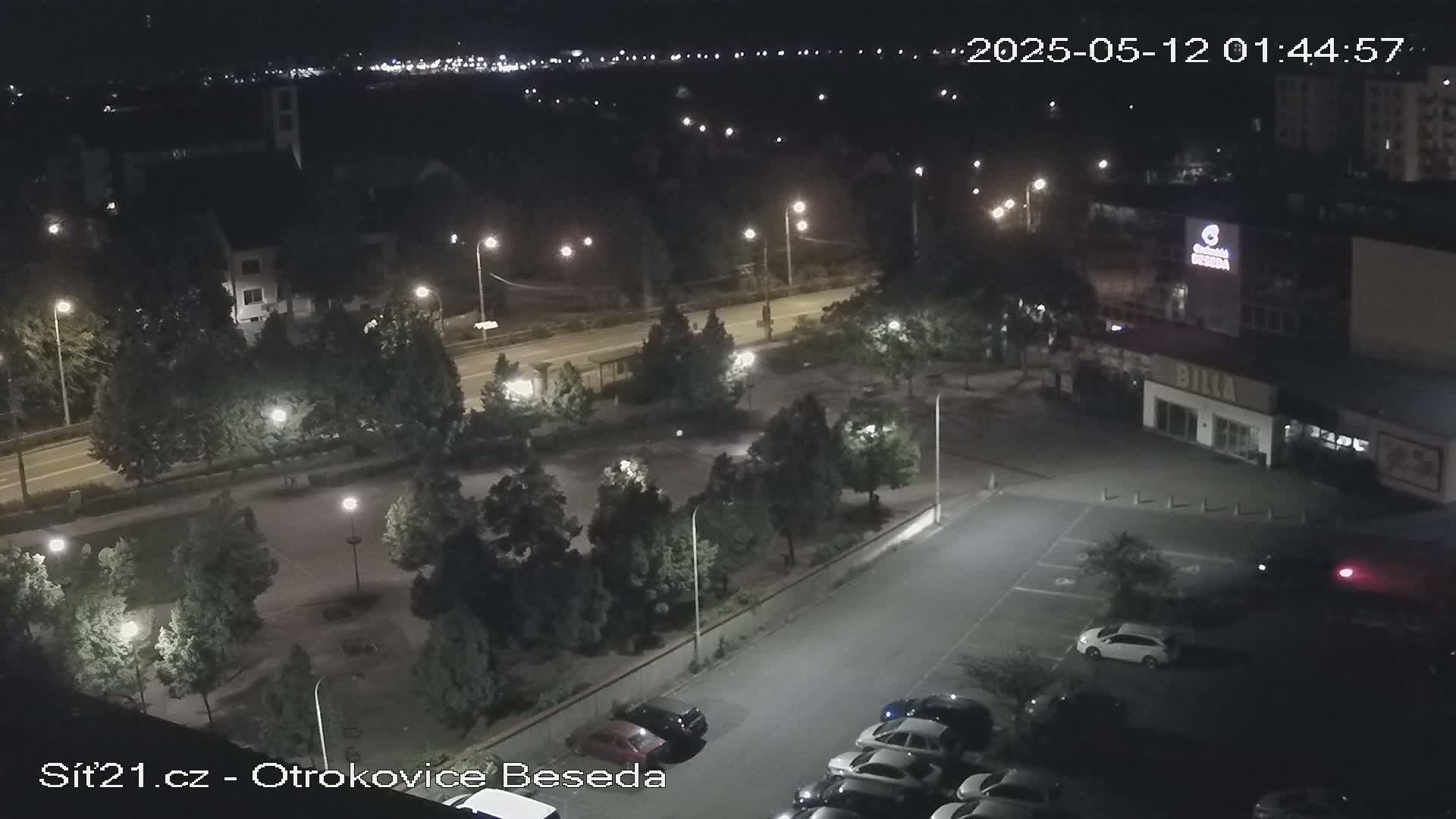 A nighttime aerial view of a parking lot and roadway, surrounded by trees and buildings, under clear skies.