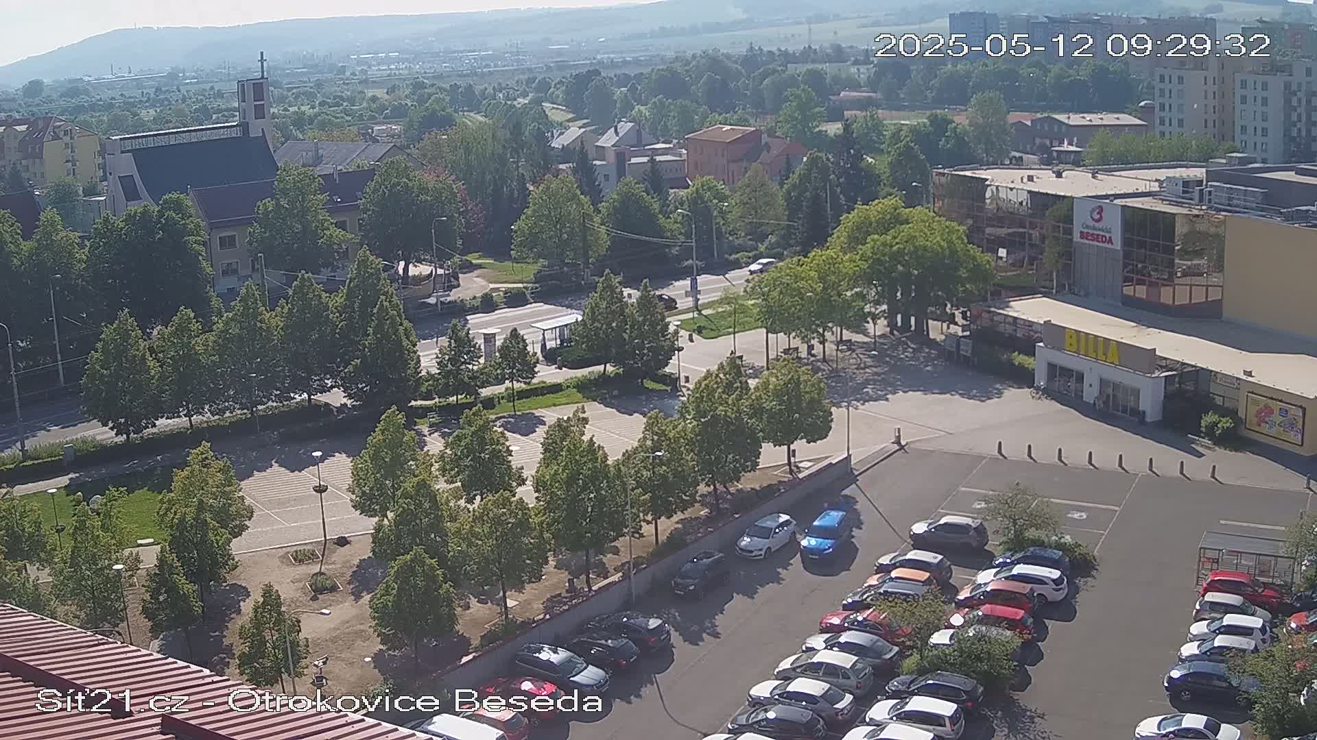 A high-angle, sunny view of a town square with a church, shops, and a parking lot filled with cars, surrounded by green trees.
