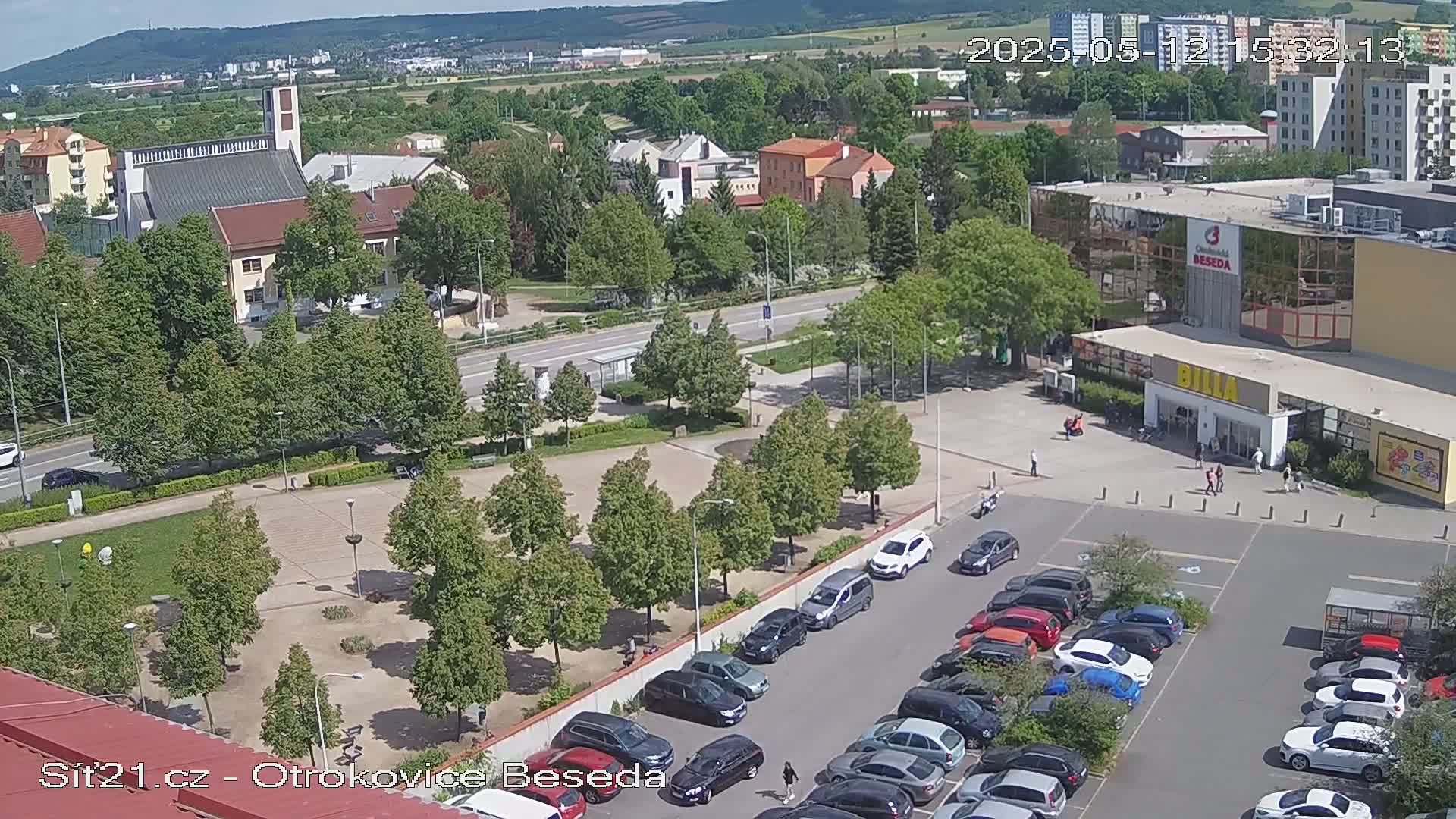 An aerial view of a town square with a parking lot, several buildings, and green spaces on a sunny day.