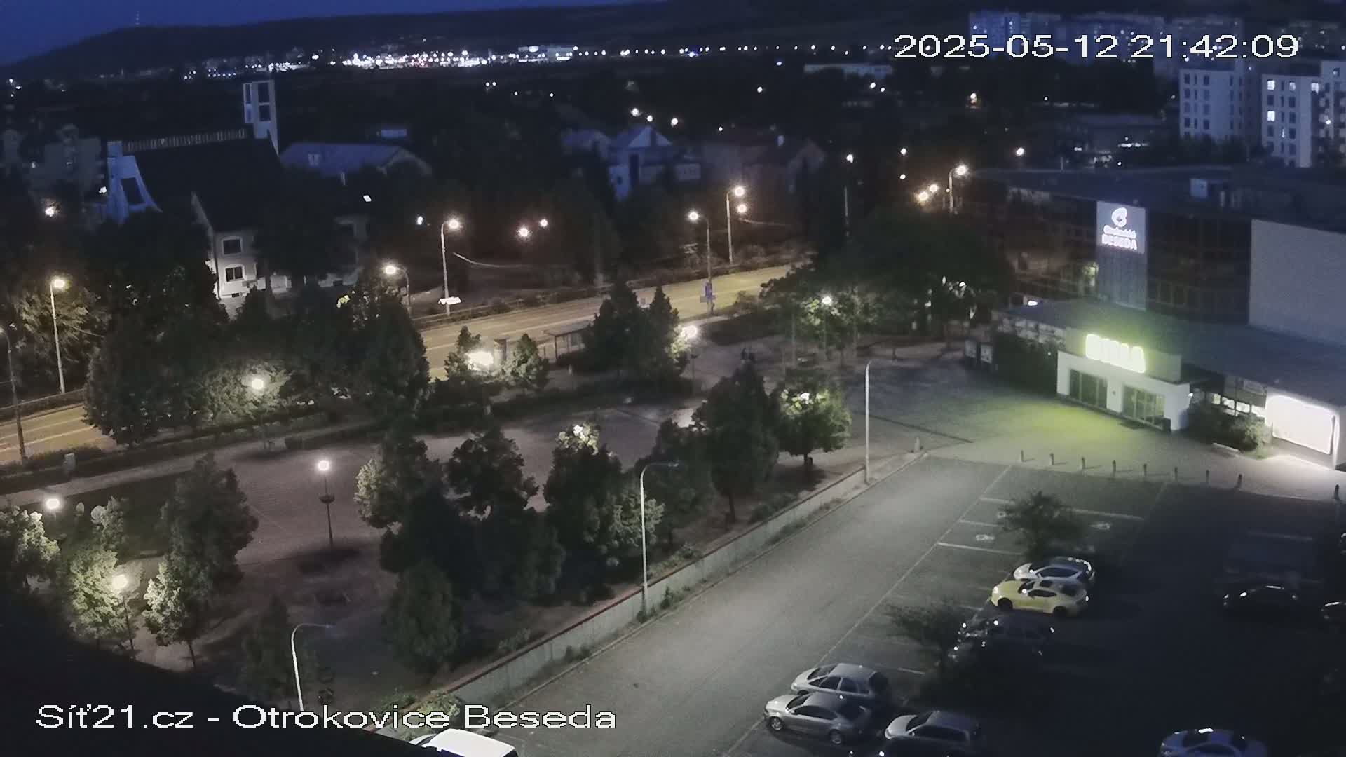 An aerial nighttime view shows a parking lot with several cars, a roadway with streetlights, and a building complex with surrounding landscaping under a dark, clear sky.