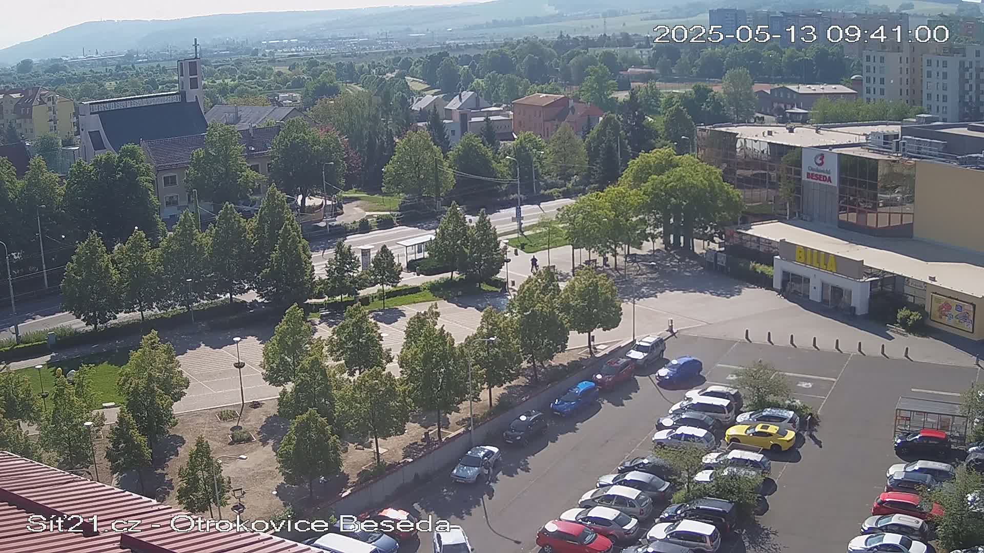 A high-angle view of a town square on a sunny day, showing a church, buildings, a parking lot with cars, and green spaces.