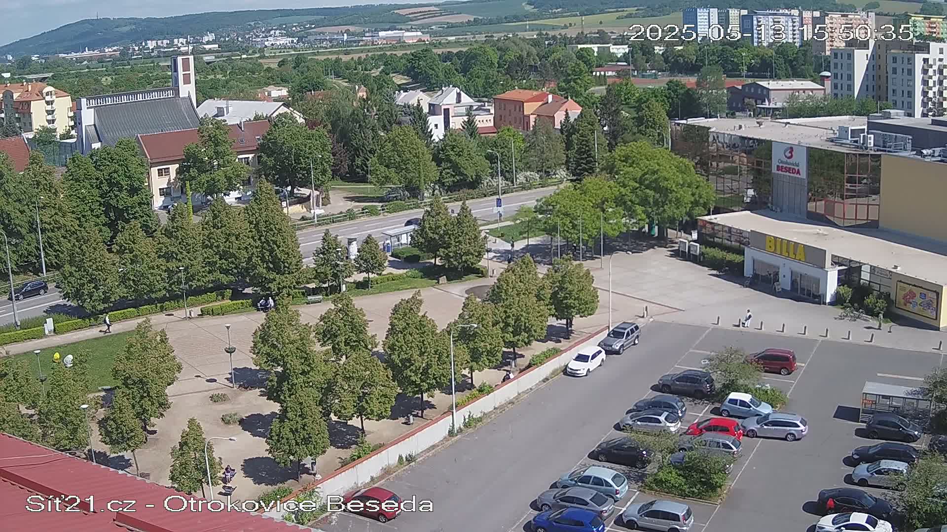 An aerial view of a town square with a church, buildings, a parking lot filled with cars, and green trees under a sunny sky.