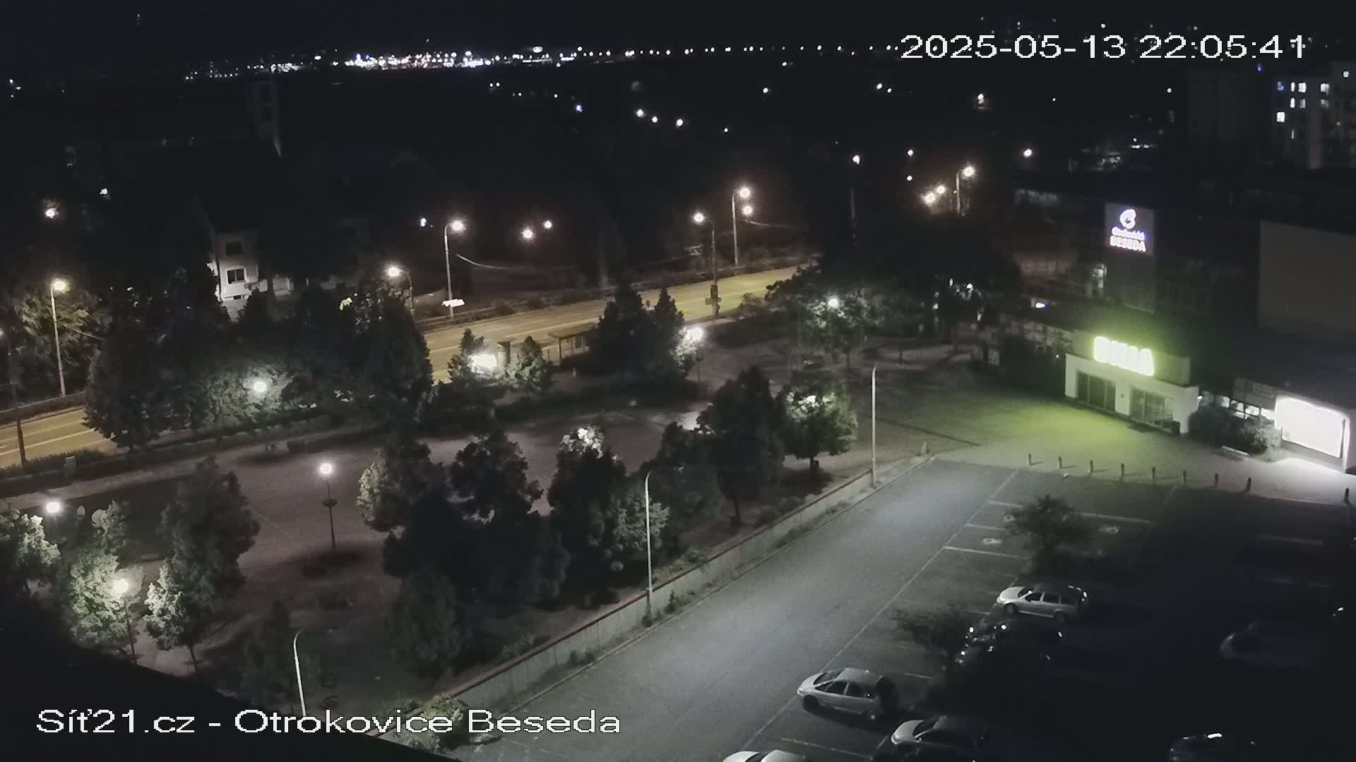 A nighttime aerial view shows a parking lot with several cars, a building, and a park-like area with trees and lights.