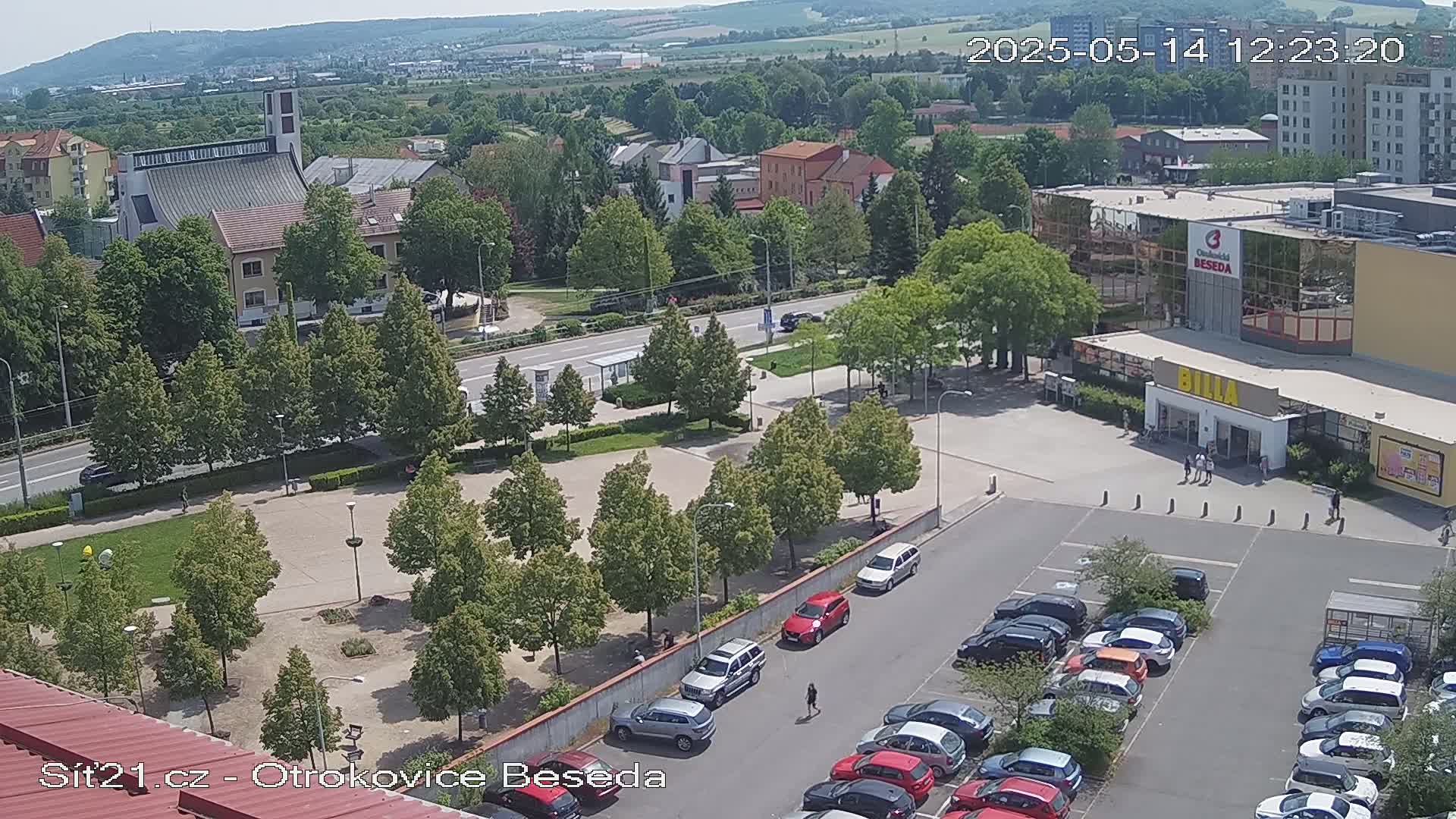 A sunny day view of a town square with a church in the background, a parking lot filled with cars in the foreground, and a row of trees lining the square.