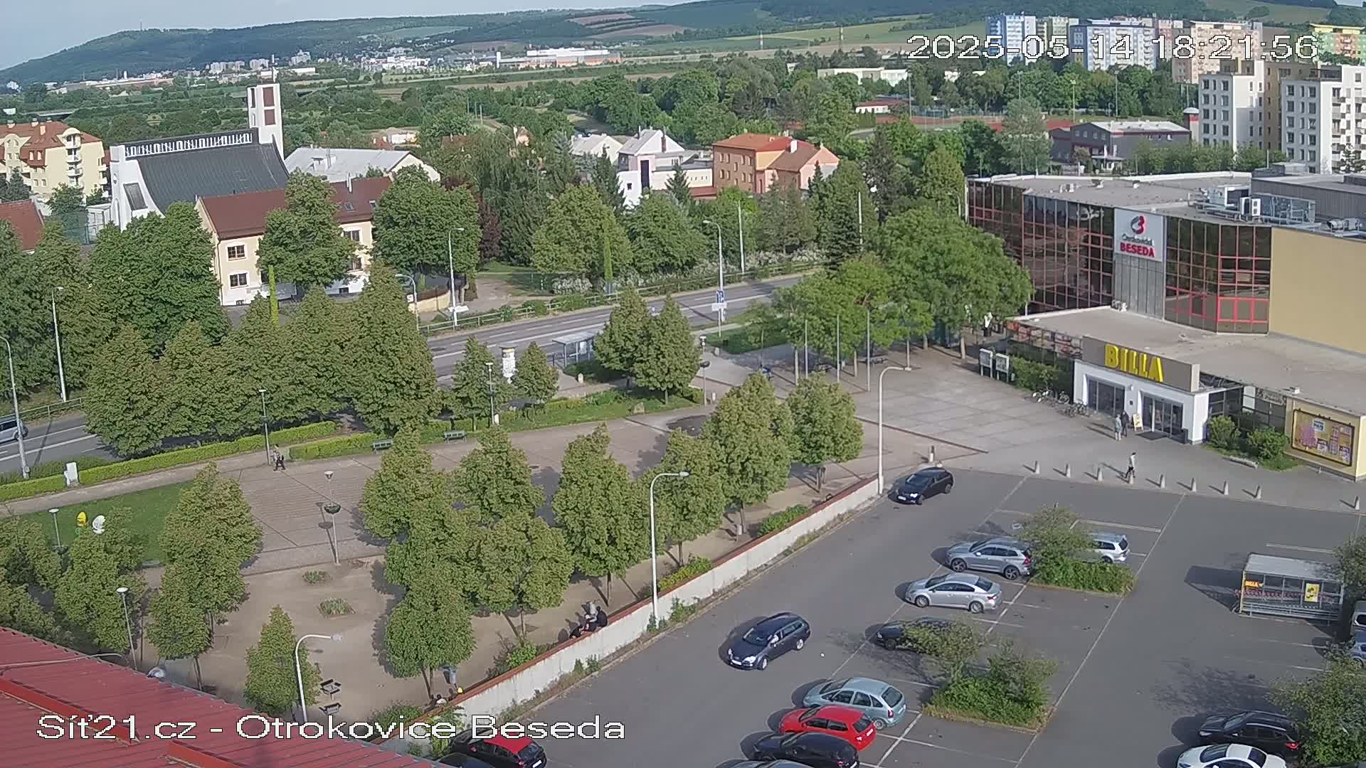 An aerial view shows a town square with a church, buildings, a parking lot with cars, and green spaces under a partly sunny sky.