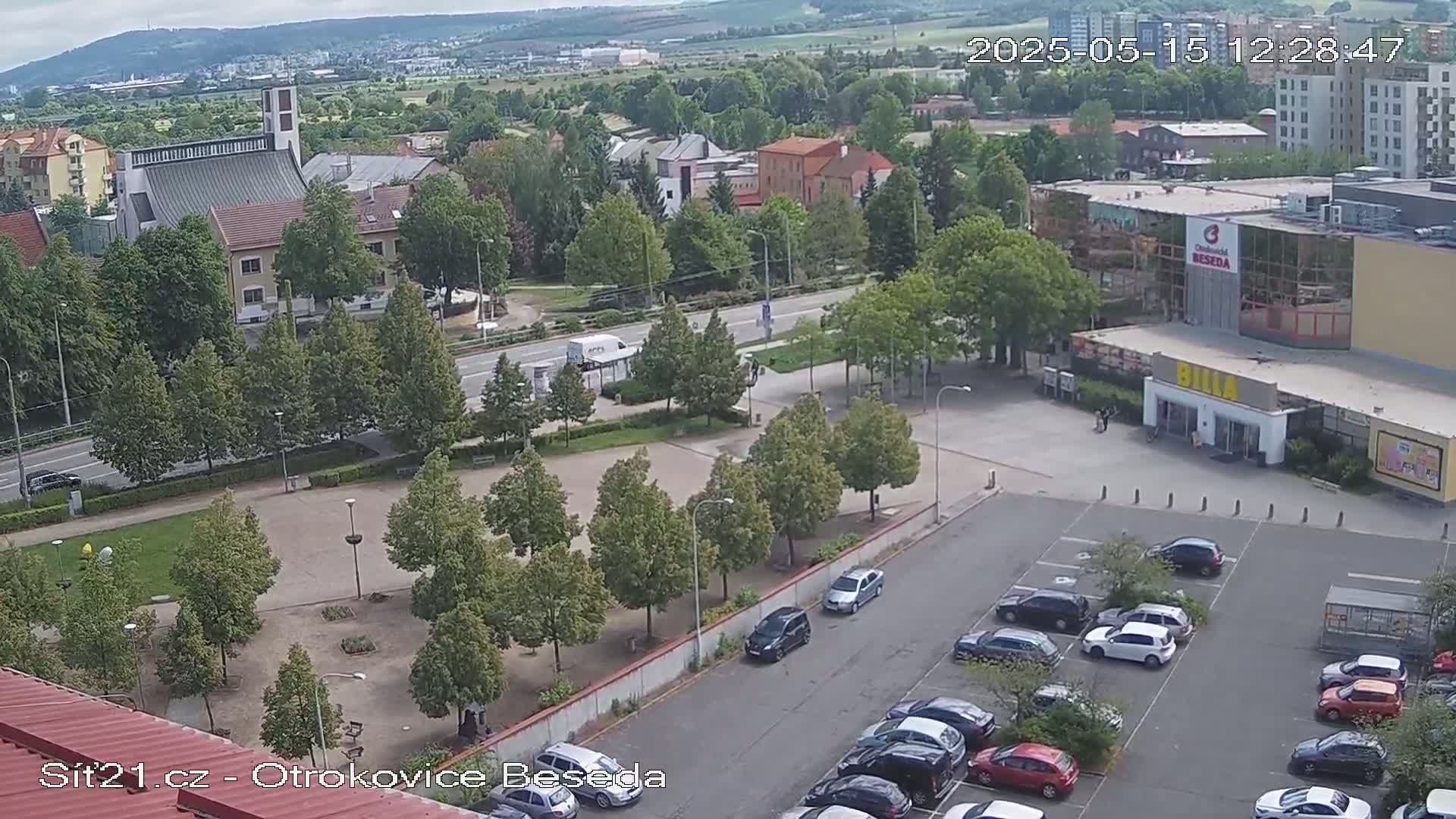 A high-angle, long shot of a town square with a church, buildings, a parking lot full of cars, and green trees under a partly cloudy sky.