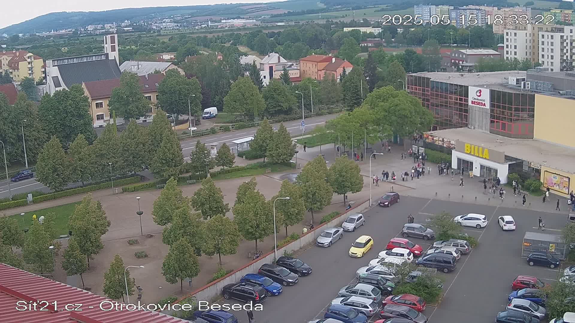 An aerial view shows a town square with a church, a large building with a parking lot full of cars, and many people walking around on an overcast day.
