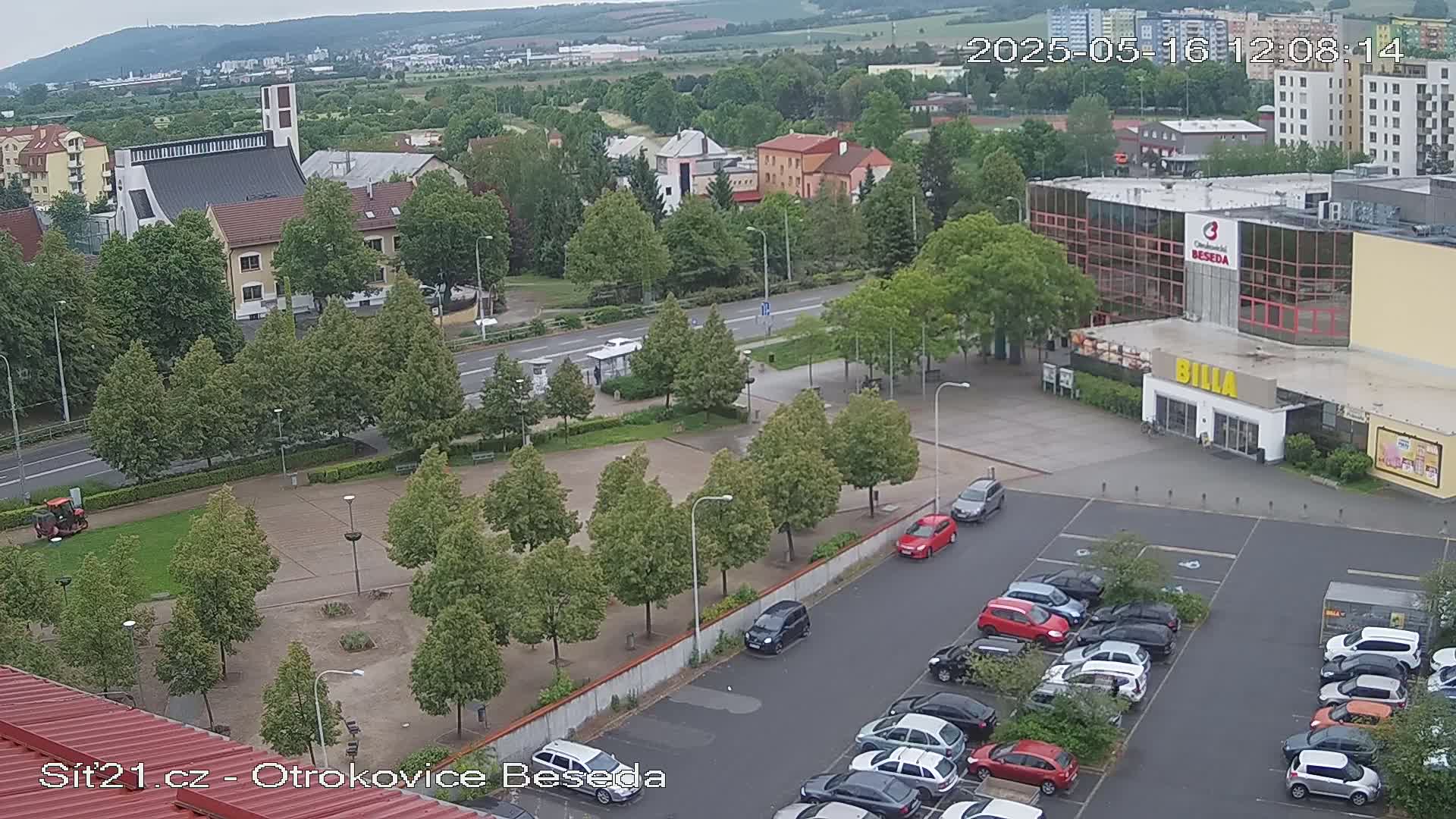 An aerial view shows a town square with a church, a large building, a parking lot full of cars, and many trees on an overcast day.