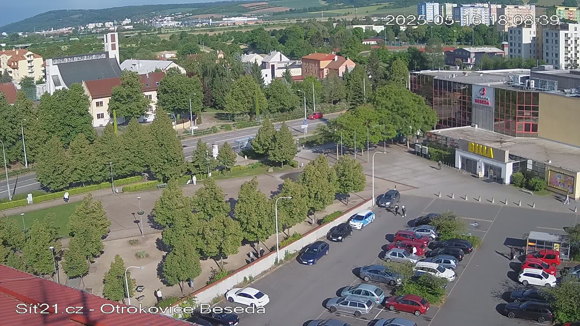 A high-angle, sunny view of a town square with a church, shops, and a parking lot filled with cars, surrounded by green trees and landscaping.