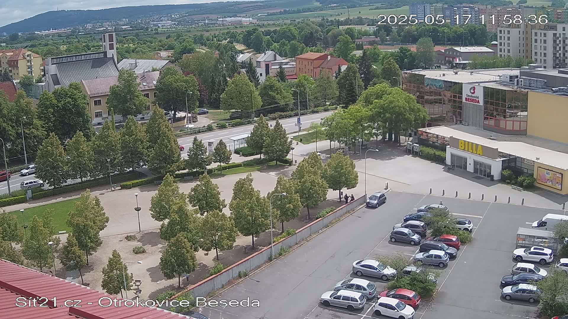 An aerial view shows a town square with green trees, a church, a large building, and a parking lot filled with cars under a partly cloudy sky.
