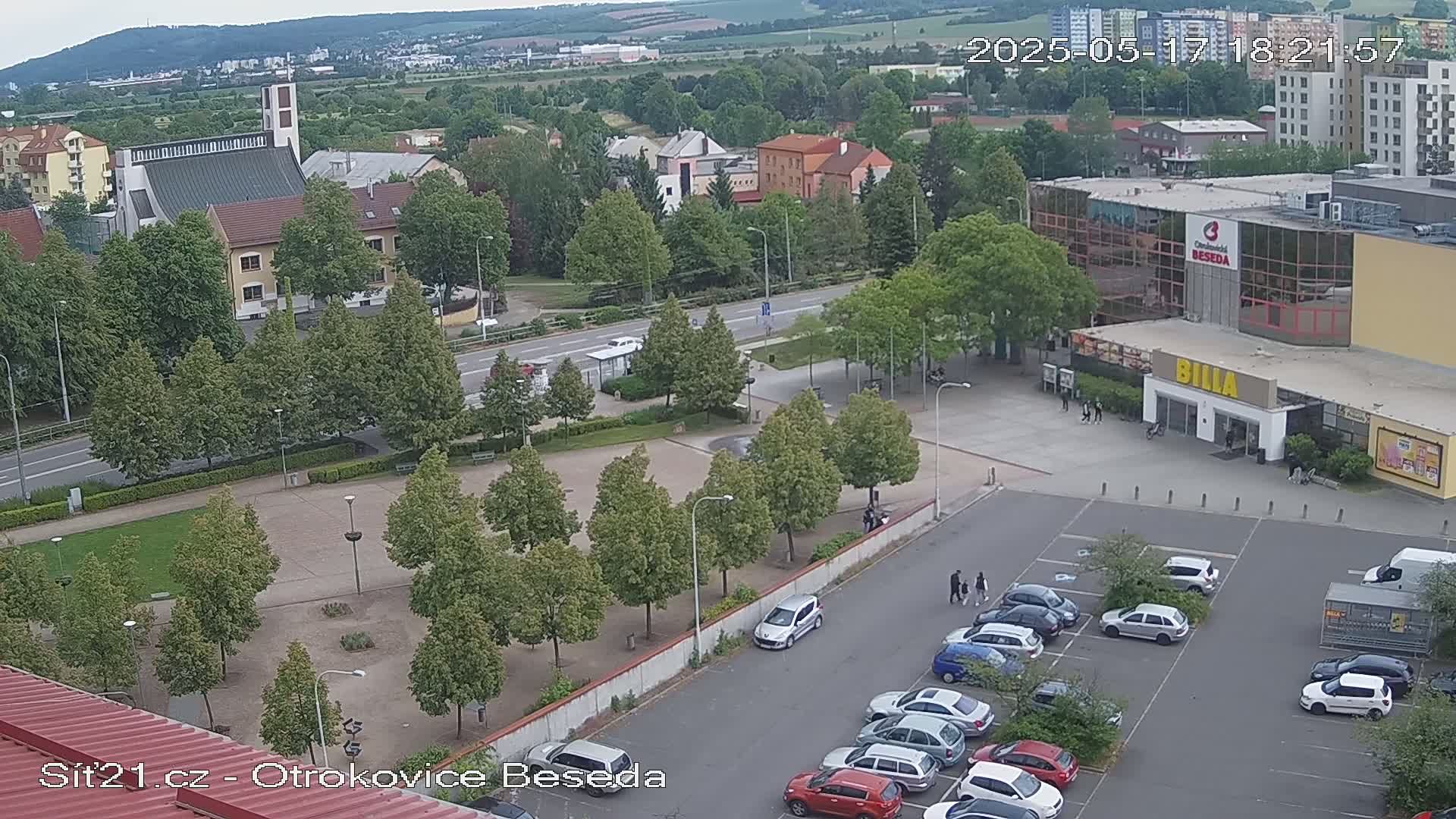 An aerial view shows a town square with a church, a large building with a parking lot, and many green trees under an overcast sky.