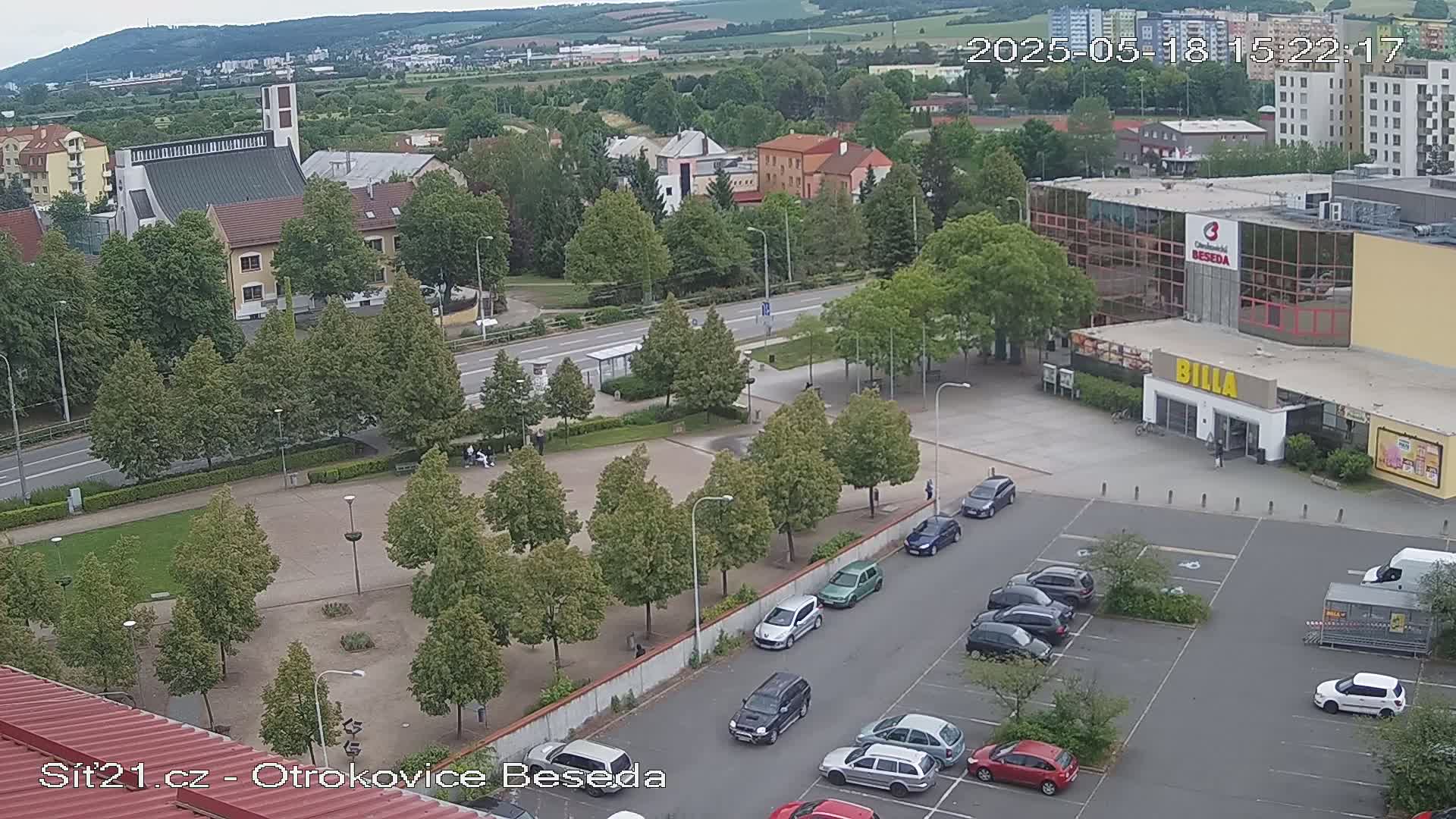 An aerial view shows a town square with a church, a supermarket, and a parking lot filled with cars, under partly cloudy skies.