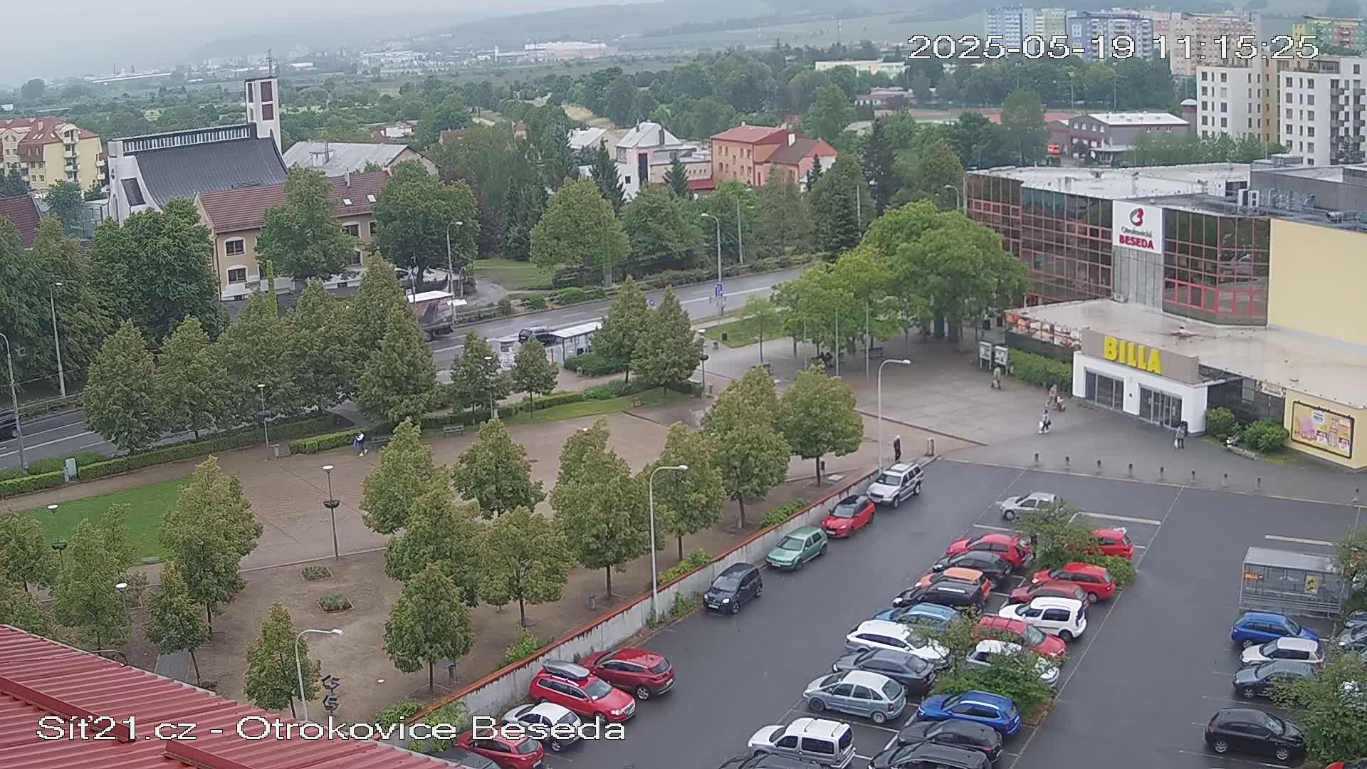 An overcast day shows a town square with a church, a large building, and a parking lot filled with cars.