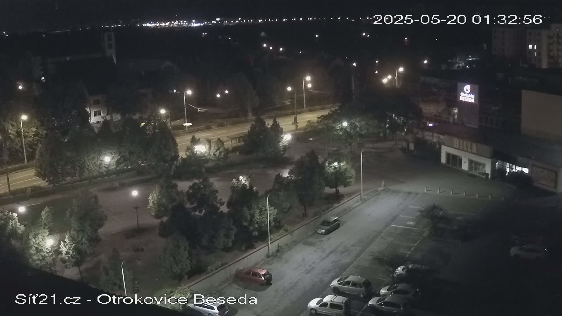 A nighttime aerial view of a parking lot with several cars, surrounded by trees and streetlights, next to a road and buildings.